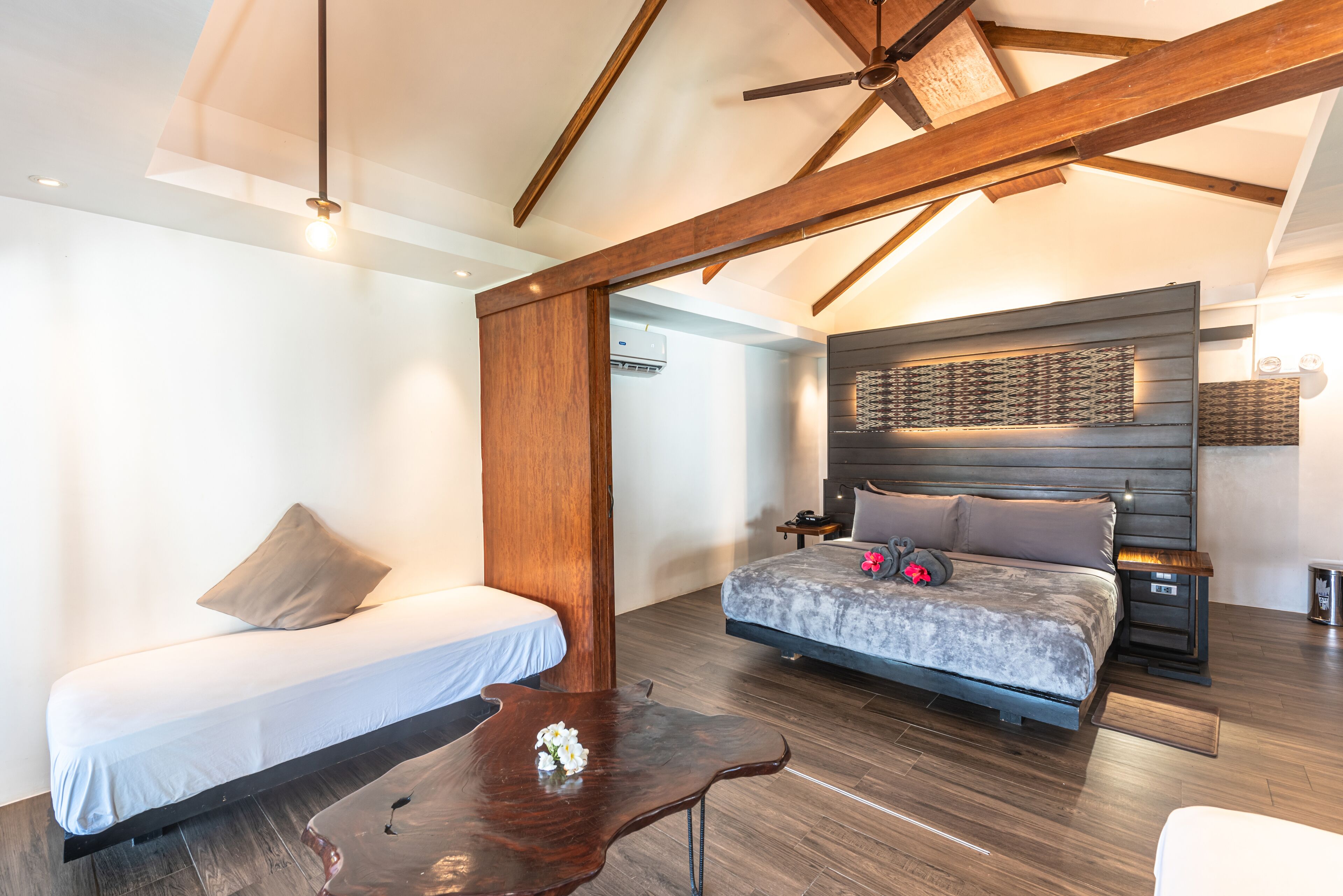 Un dormitorio moderno con una cama grande, una cama más pequeña, vigas de madera, un ventilador de techo y una mesita de noche con lámpara.