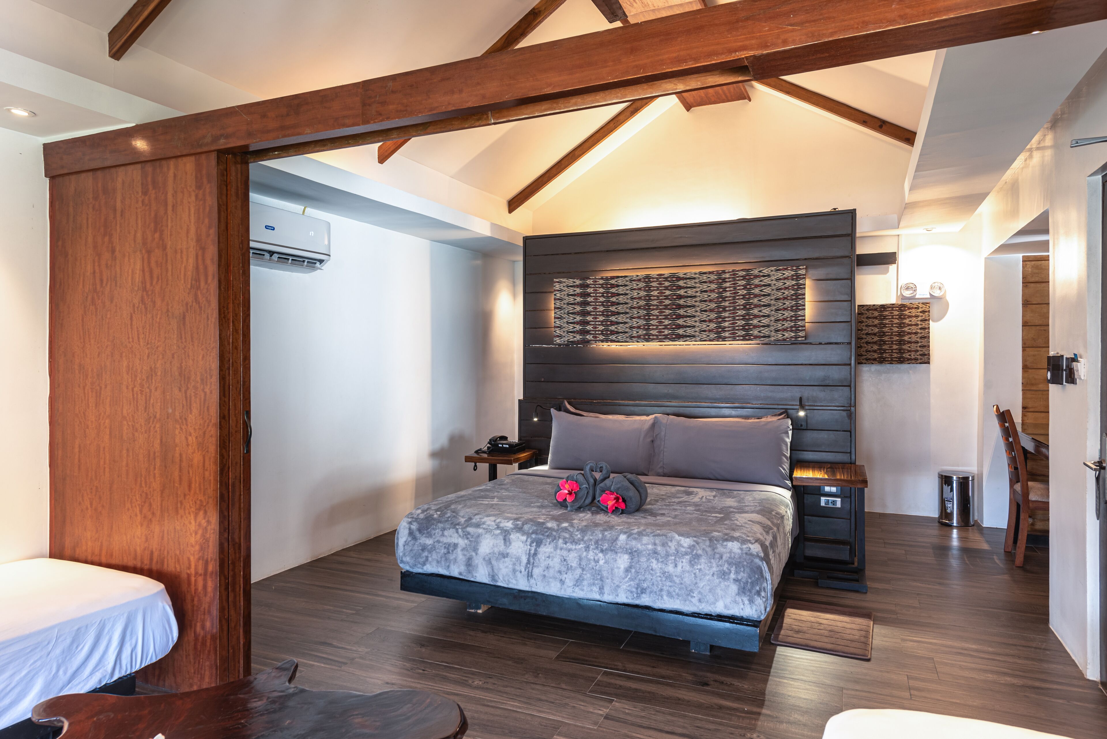 A bedroom with a large bed, wooden beams, a headboard with a patterned design, and a bedside table with a lamp.