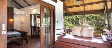 Eine Holzterrasse mit einer Bank und Kissen, einem Deckenventilator und Blick auf Grünflächen.