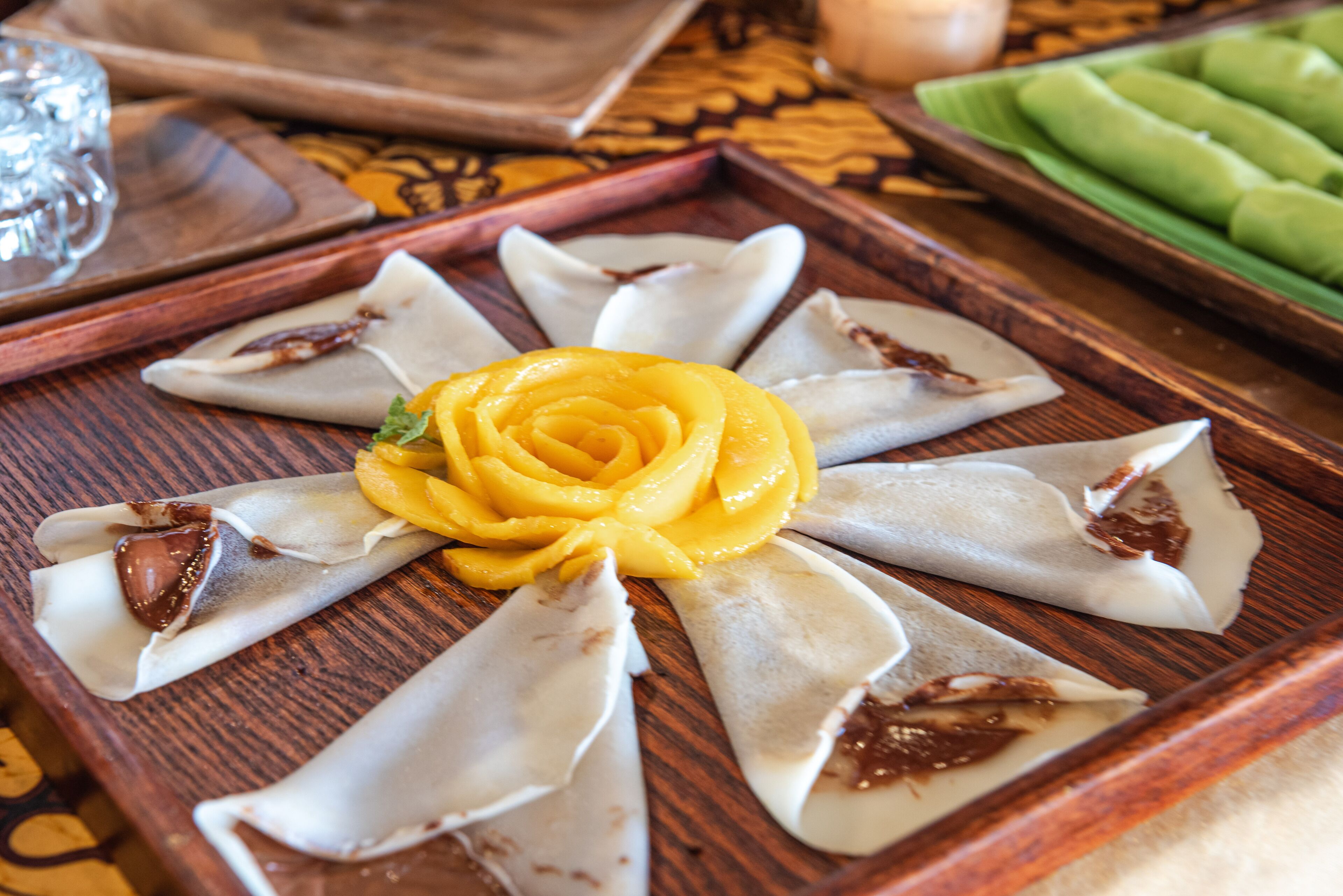 A wooden tray with a decorative arrangement of food items, including a yellow flower-like garnish and several wrapped items.