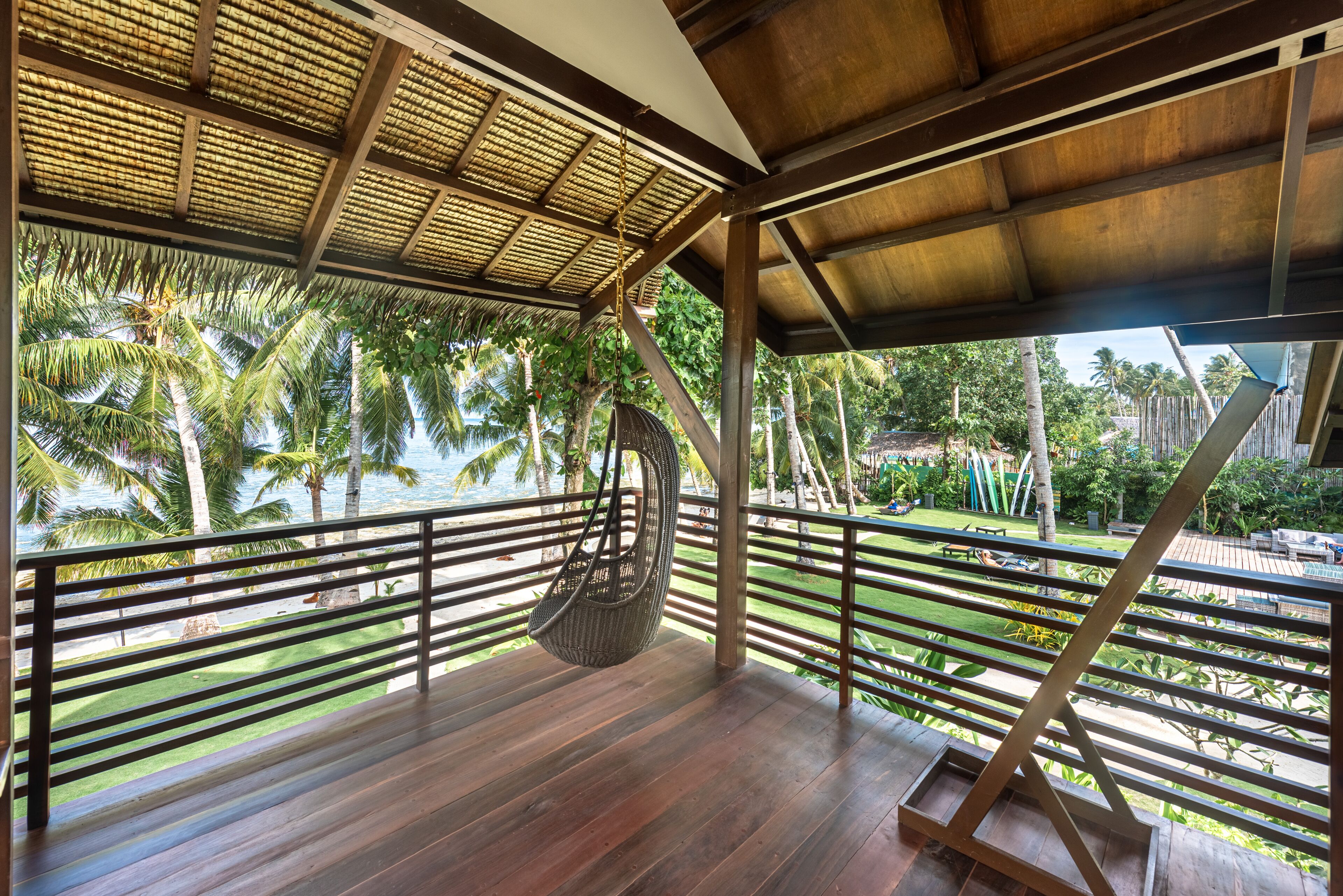 A wooden deck with a hanging chair, a railing, and a view of palm trees and a beach.