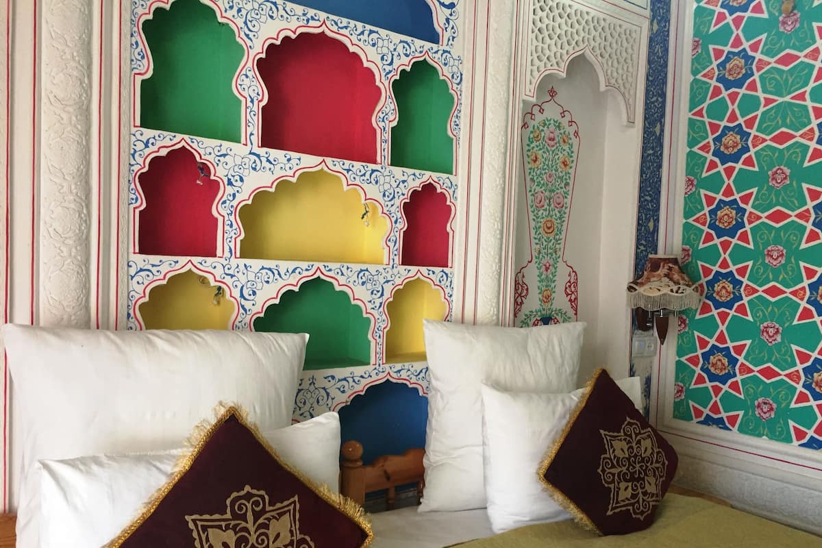 Une chambre au décor traditionnel, avec un lit agrémenté d’oreillers blancs et bordeaux, un mur aux couleurs vives orné de motifs géométriques et un plafond richement décoré.