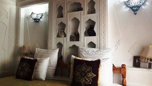 Een kamer met een bed, decoratieve kussens, een wandplank met kleine objecten en een wandlamp.