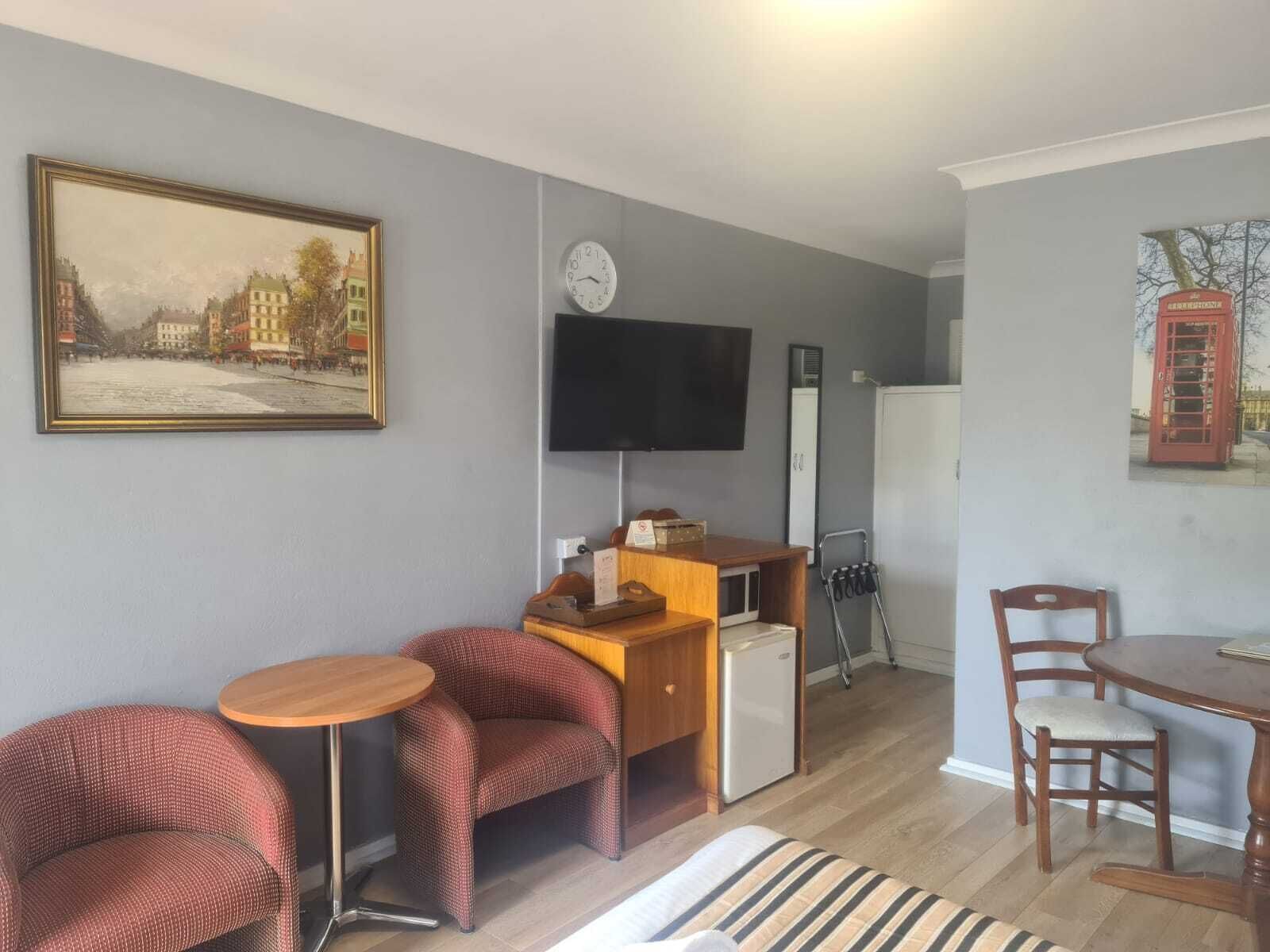 A compact living space with a sofa, two armchairs, a small wooden table, a television, a refrigerator, and a painting of a street scene.