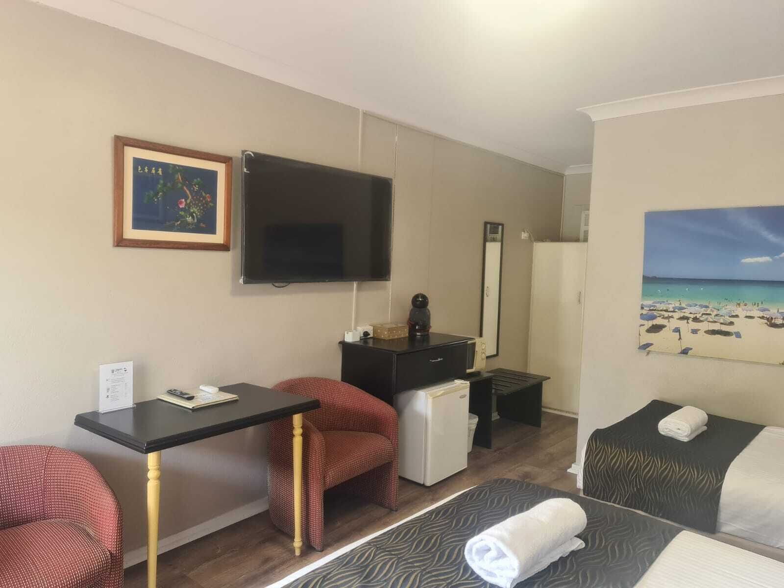 A hotel room with a bed, a desk with a chair, a television, a refrigerator, and a painting of a beach.