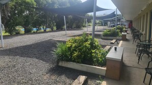 An outdoor seating area with tables and chairs under a shaded canopy, surrounded by gravel and plants. - Valley View Motel (Murrurundi)