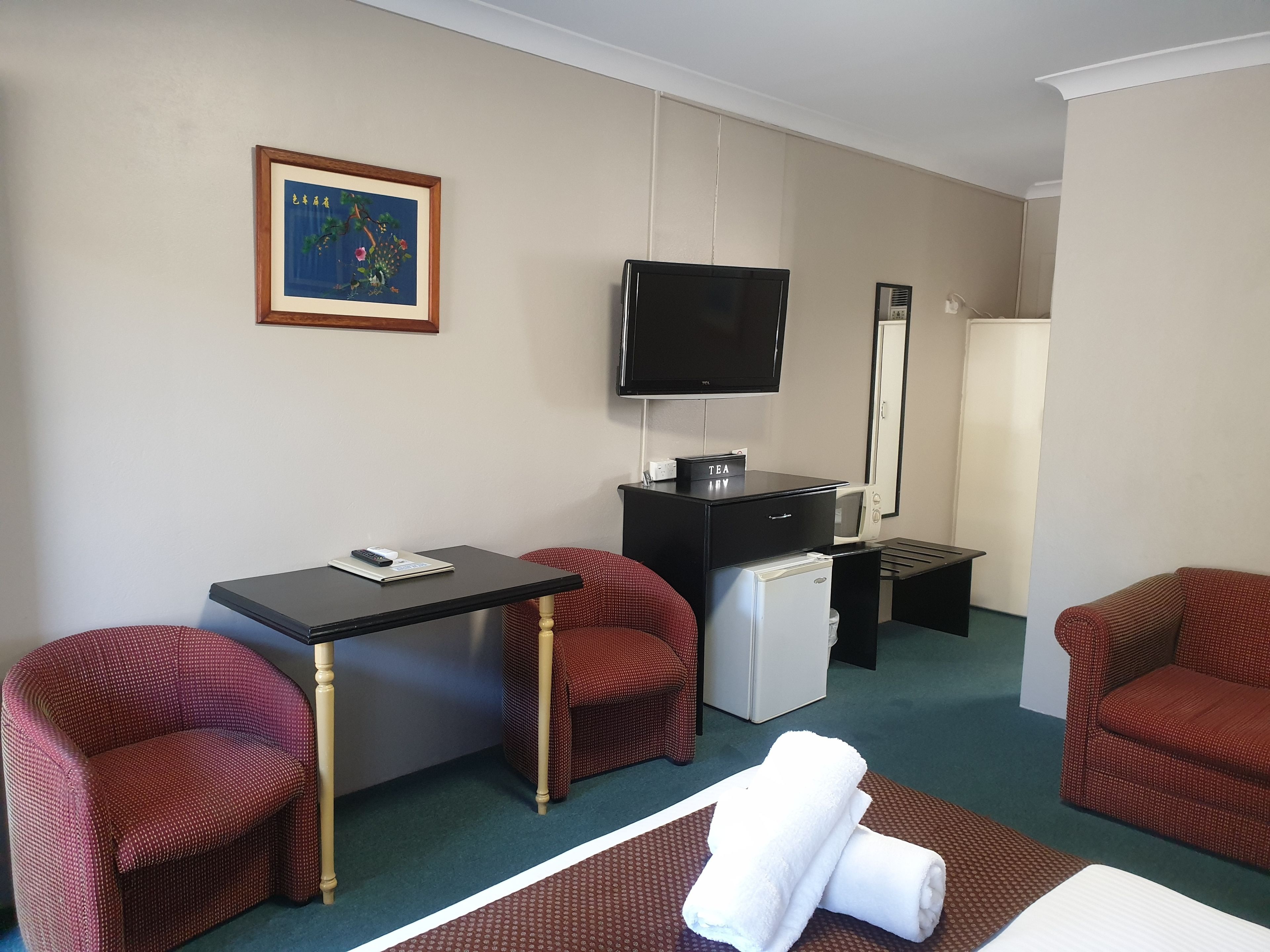 Quarto de hotel com cama, escrivaninha, televisão, frigorífico e duas poltronas vermelhas.