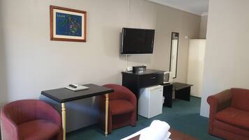 Quarto de hotel com cama, escrivaninha, televisão, frigorífico e duas poltronas vermelhas.