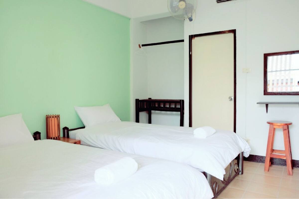 A room with two beds, a wooden headboard, a small stool, and a fan.