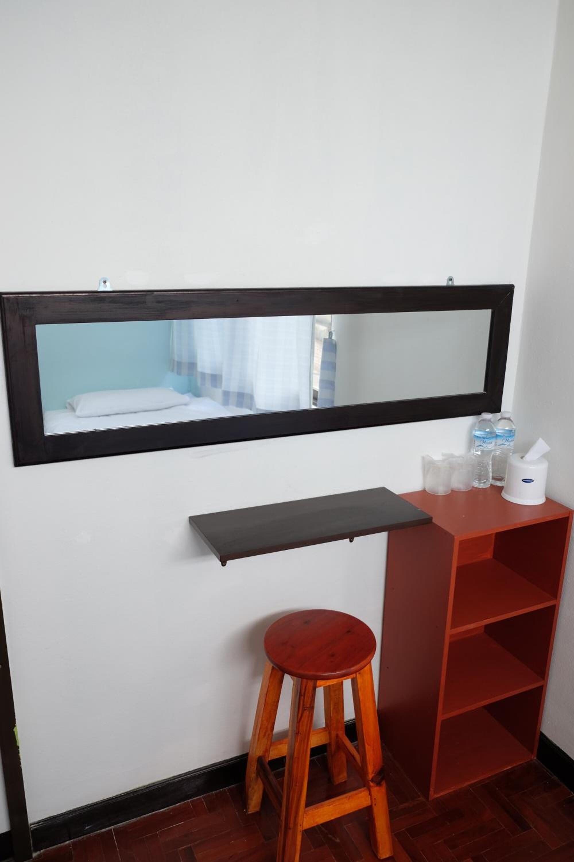 A small room with a bed, a mirror, a stool, and a shelf with toiletries.