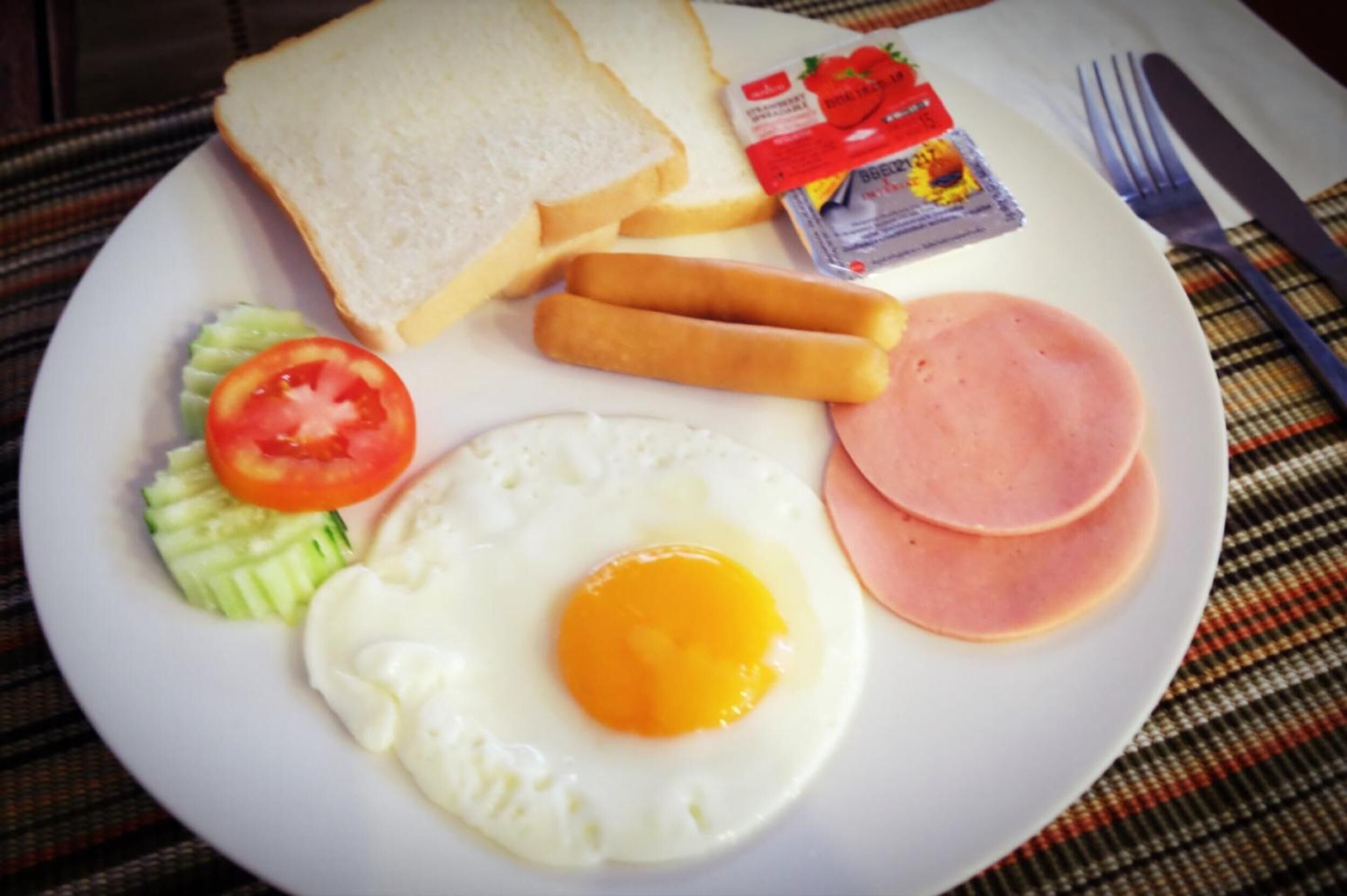 A plate with a fried egg, ham, tomato, cucumber, bread, and cheese.
