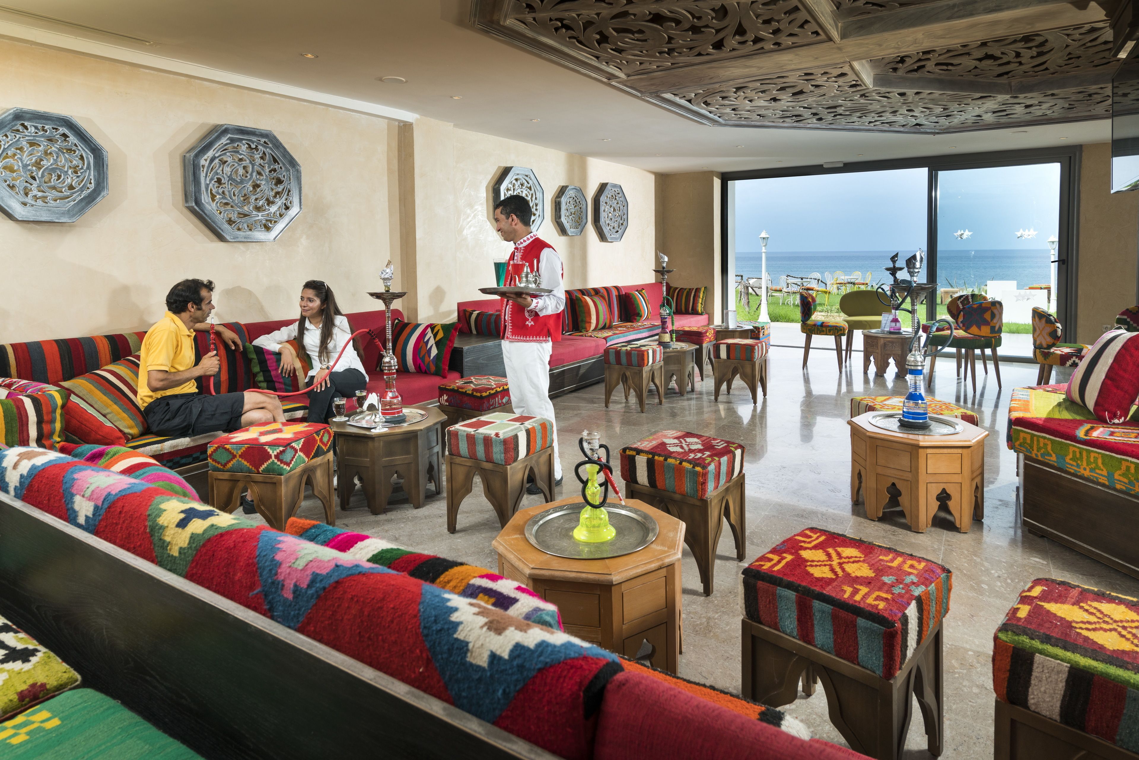 Un vaste espace salon avec des sièges aux couleurs vives, un serveur qui apporte les boissons et une vue sur la mer grâce à de grandes fenêtres.