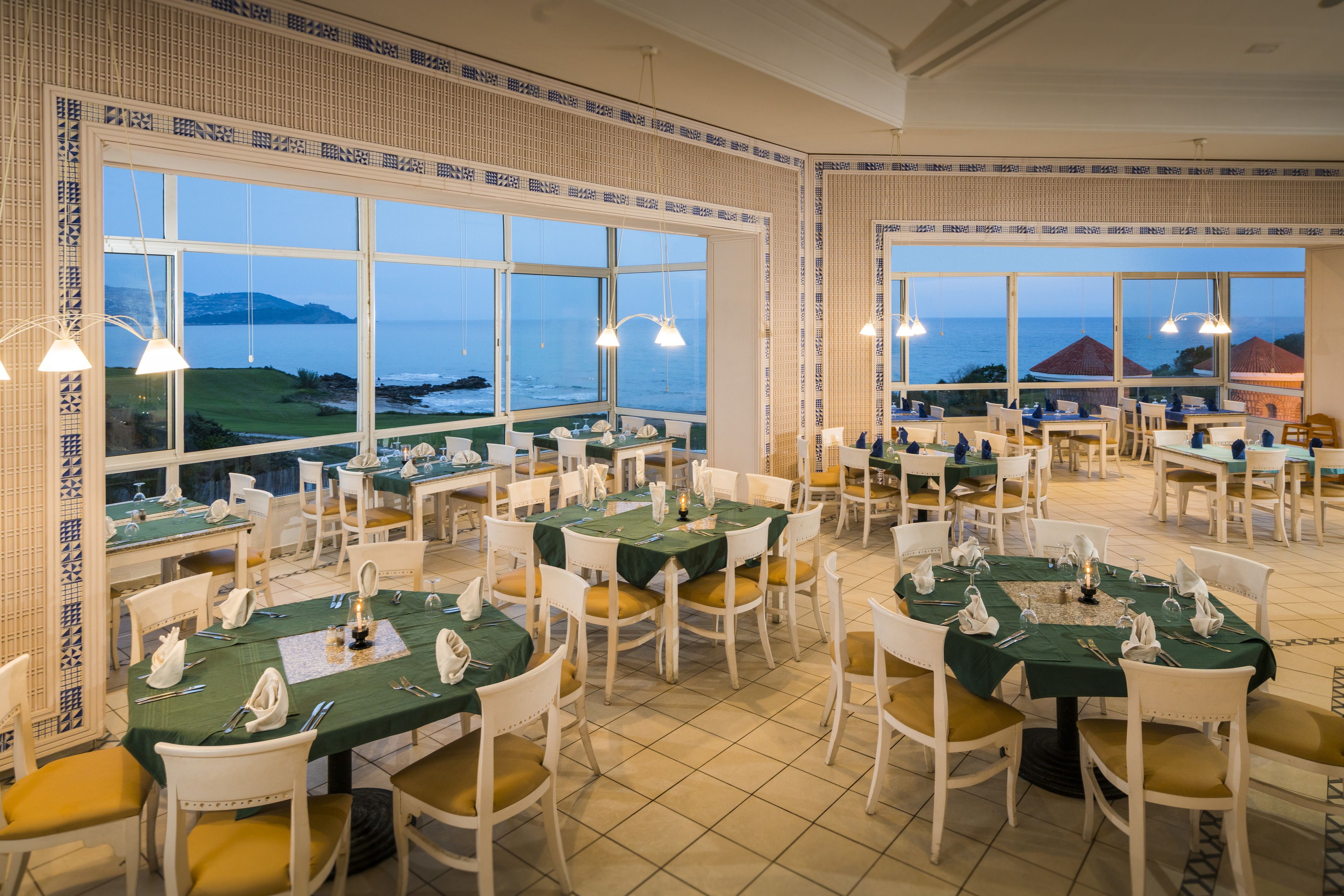 Un restaurante con varias mesas dispuestas para comer, amplias ventanas con vista al mar y decoración de pared con diseño.