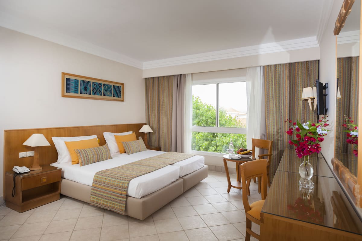 Une chambre d’hôtel comprenant un lit, une table de chevet, une lampe, une chaise, une table à manger, un vase de fleurs et un tableau au mur.