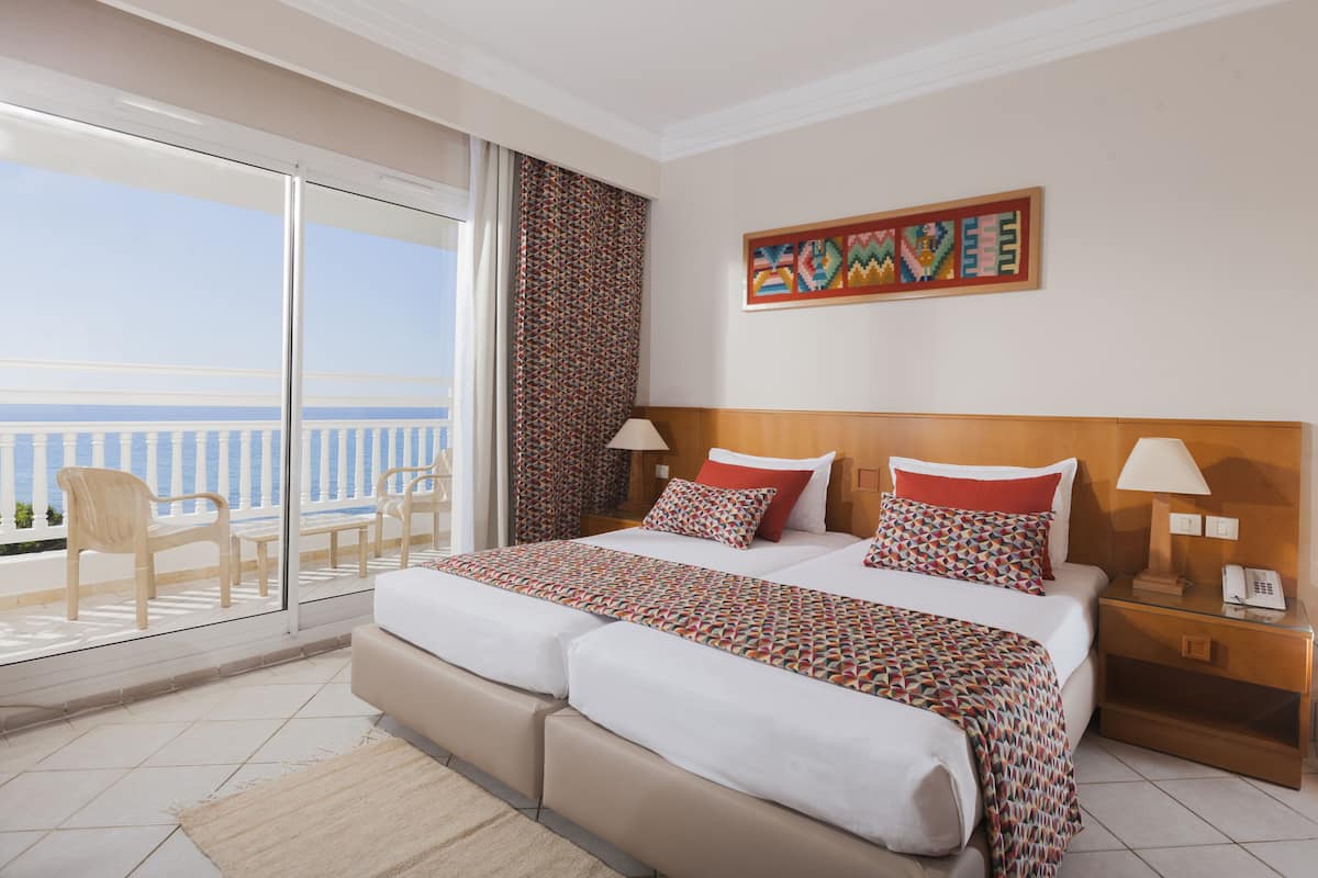 Une chambre d’hôtel avec un grand lit, deux tables de chevet, une petite table, une chaise, un balcon avec vue sur la mer et une œuvre d’art murale aux couleurs vives.