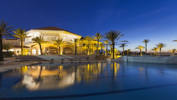 Un hotel grande y moderno con piscina, palmeras y una fachada bien iluminada.