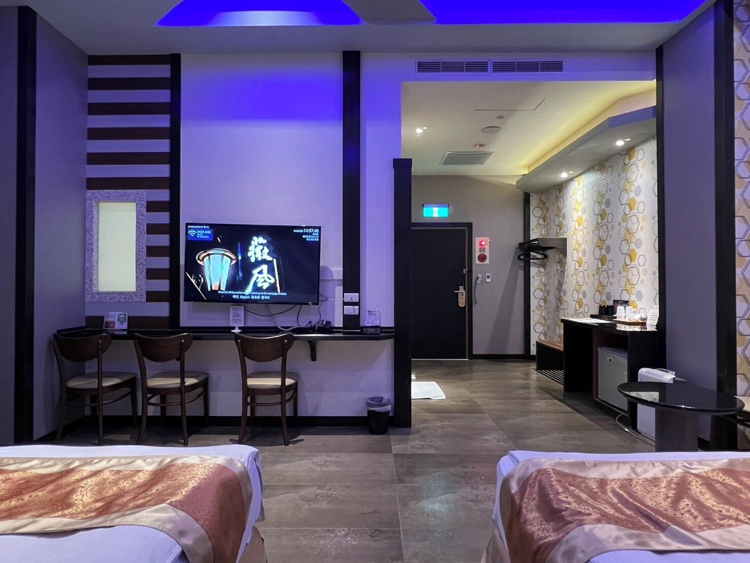 Quarto de hotel com televisão, duas camas, uma escrivaninha com cadeiras e banheiro com espelho e pia.