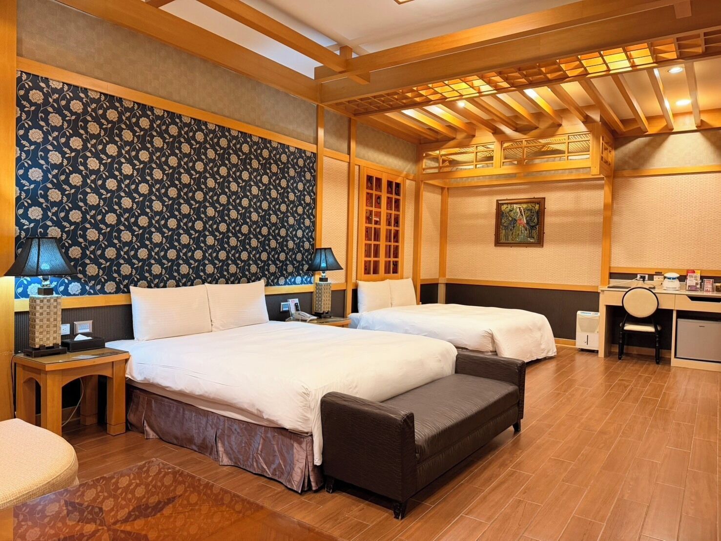 A hotel room with two beds, a desk, a chair, a lamp, and a patterned wallpaper.