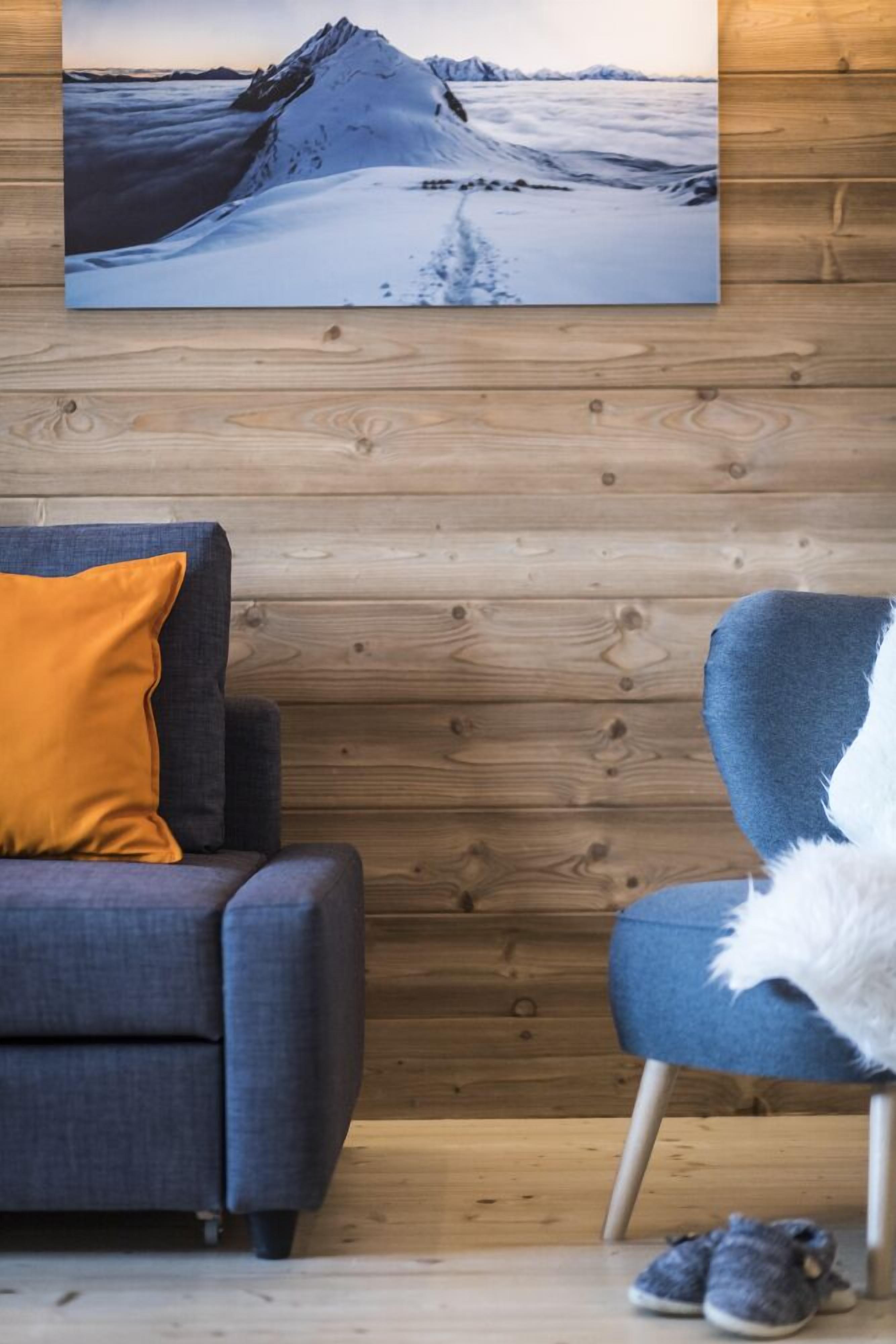 A room with a wooden wall, a grey sofa with an orange pillow, a blue chair with a white fluffy pillow, and a pair of shoes on the floor.