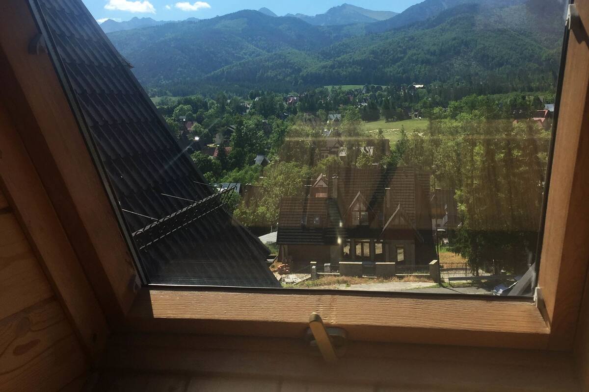 Une vue depuis une fenêtre à cadre en bois, donnant sur une vallée avec des maisons et des montagnes.