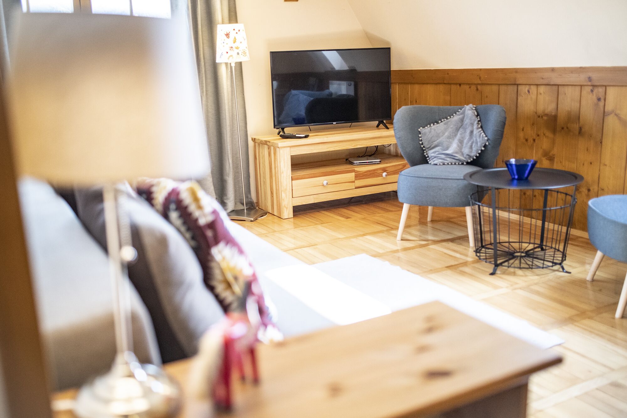 Ein gemütliches Wohnzimmer mit Holzboden, einem Fernseher auf einem Holzbodenständer, einem grauen Sessel, einem kleinen runden Tisch und einer Lampe.