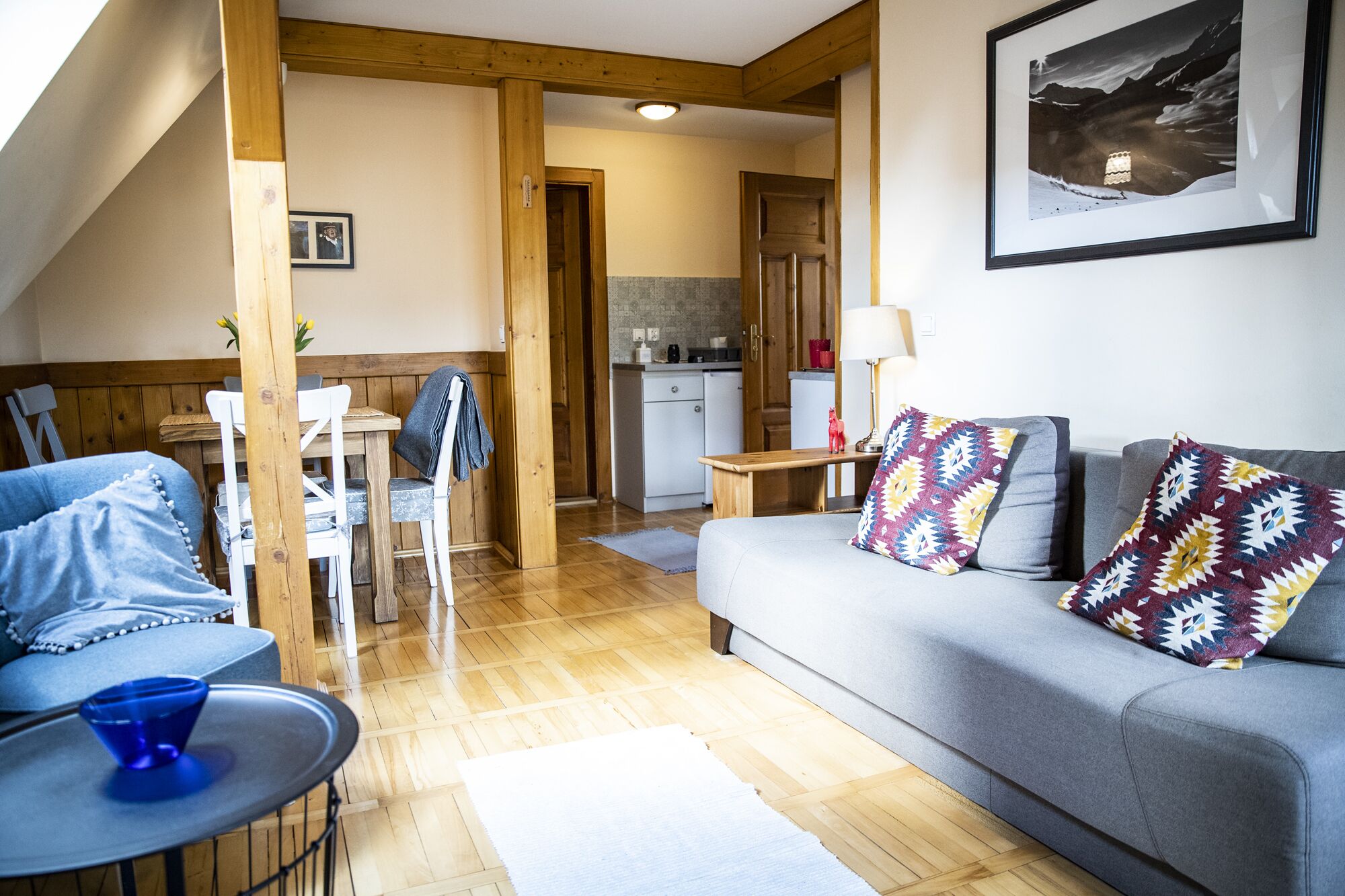 Un salon chaleureux avec un canapé gris, un fauteuil bleu et une petite table sur laquelle se trouve un bol bleu. On aperçoit, au fond, une table à manger en bois avec des chaises, et une cuisine est visible à travers une porte ouverte.