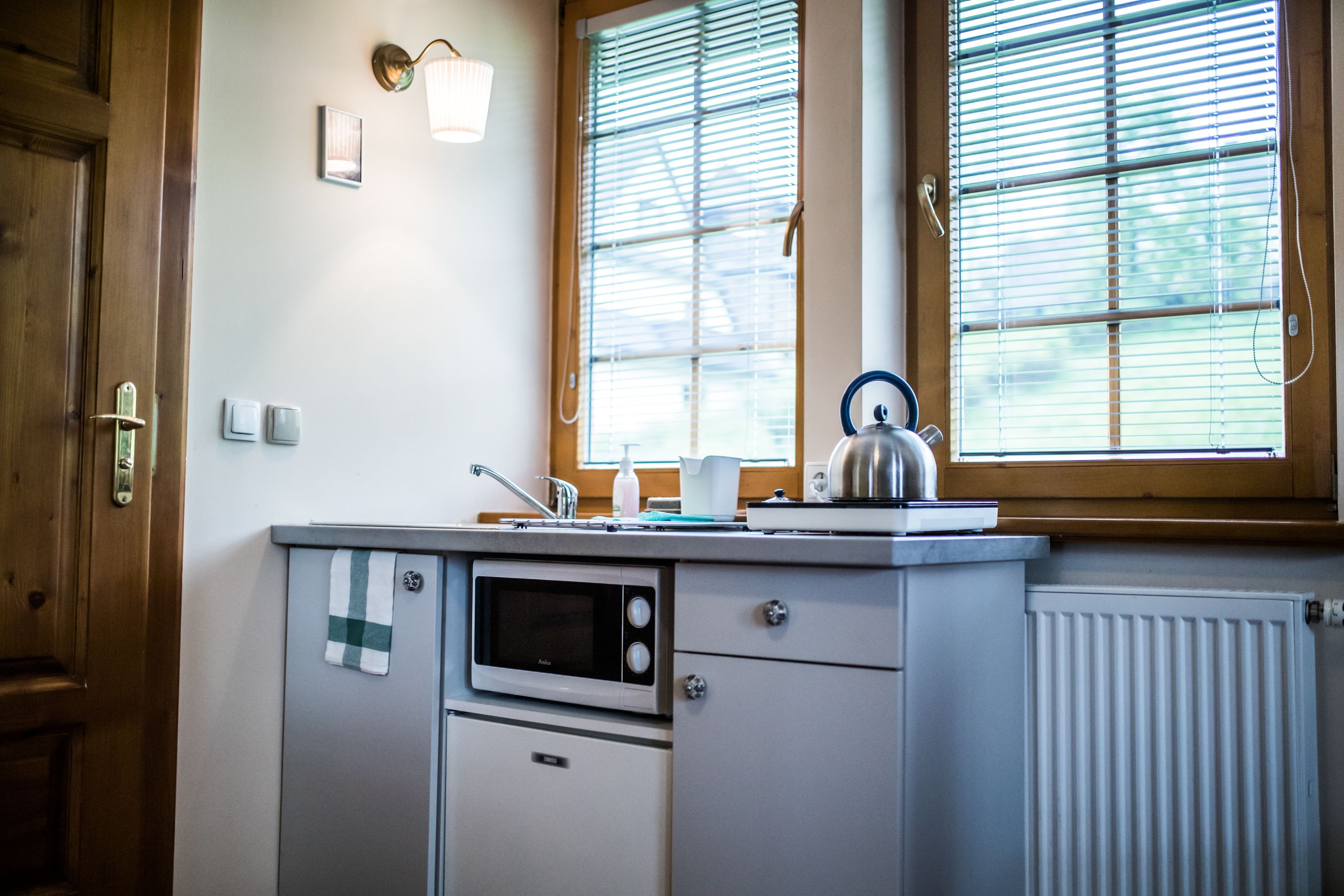 A compact kitchen with a microwave, kettle, and a window with blinds.