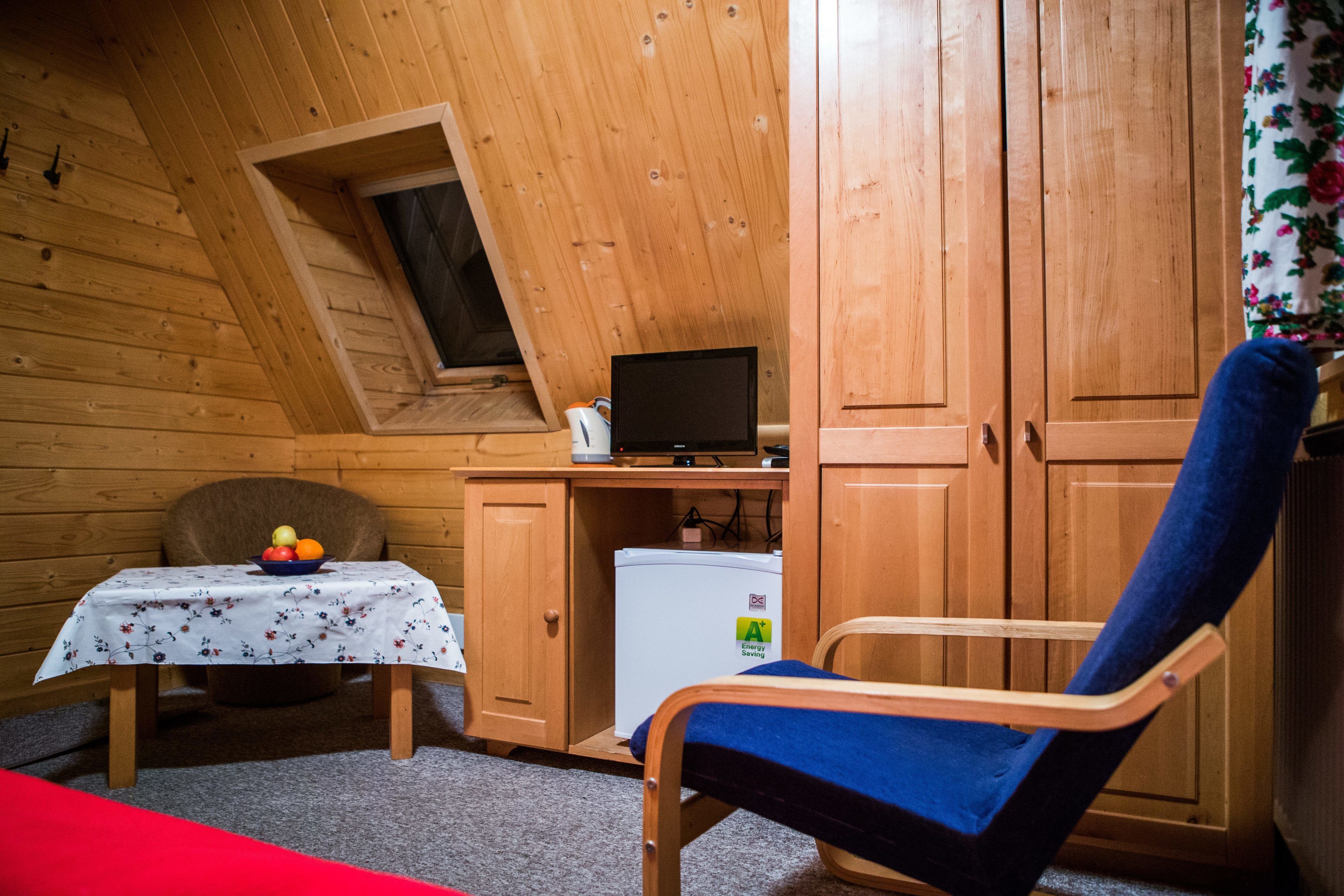 Une petite pièce avec un plafond en bois, un bureau avec un ordinateur, une chaise, une table avec un panier de fruits et un réfrigérateur.