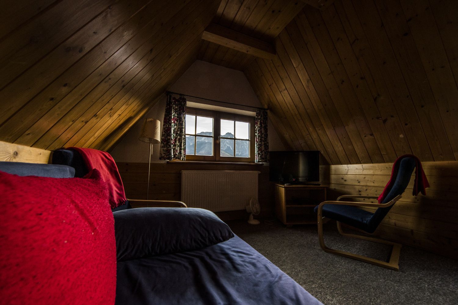 A cozy attic bedroom with a sloped ceiling, a window with curtains, a bed with red and blue bedding, a wooden chair, and a television.