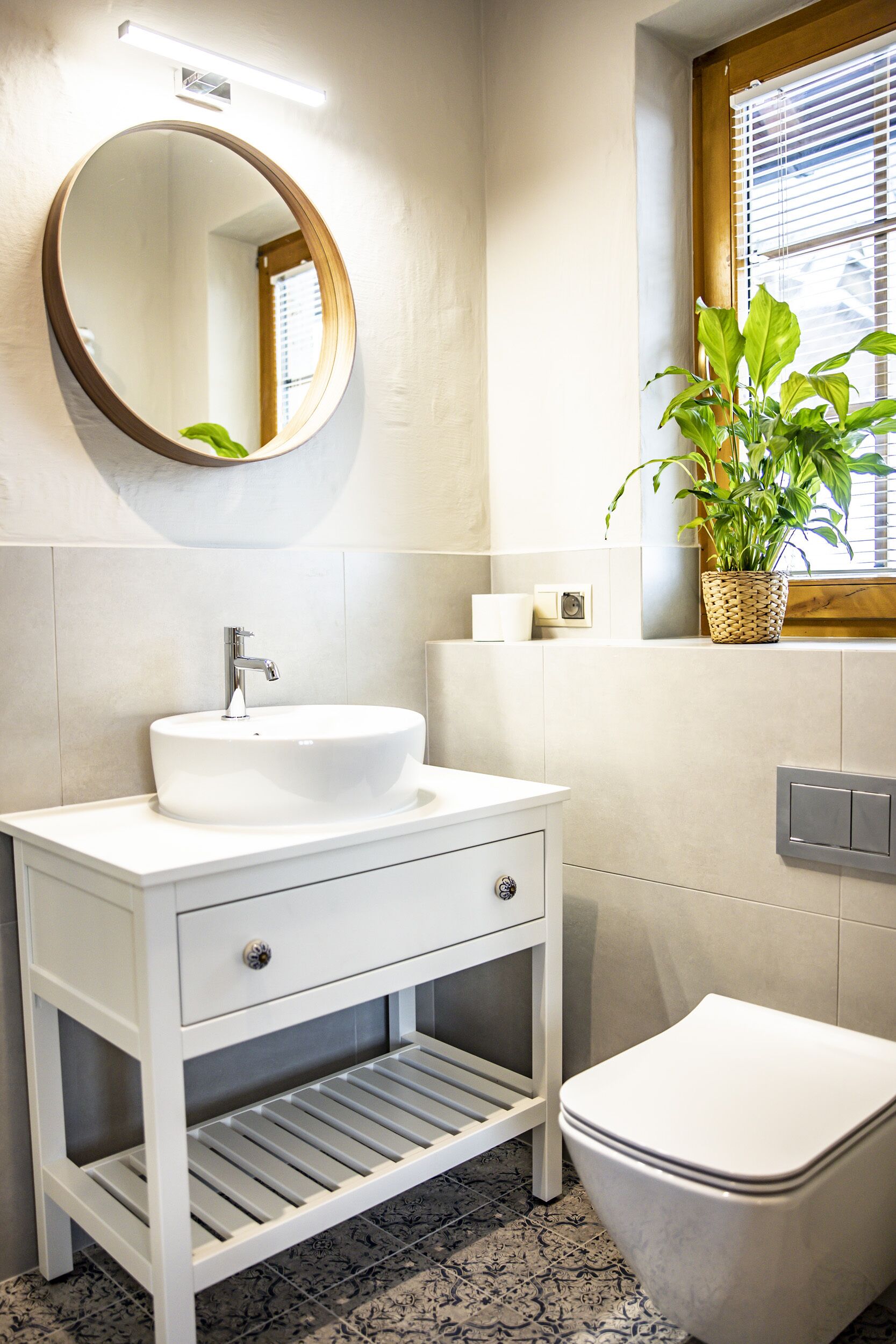 A bathroom with a white vanity, a round mirror, a white sink, a toilet, and a potted plant on the windowsill.