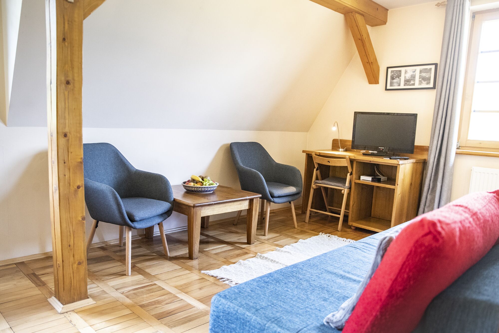 A cozy room with two blue armchairs, a small wooden table with a bowl of fruit, a desk with a computer and TV, and a bed with a red pillow.