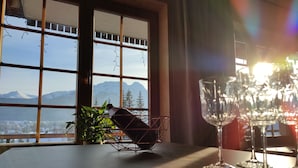 Uma mesa com uma garrafa, copos de vinho, uma planta e vista das montanhas pela janela.