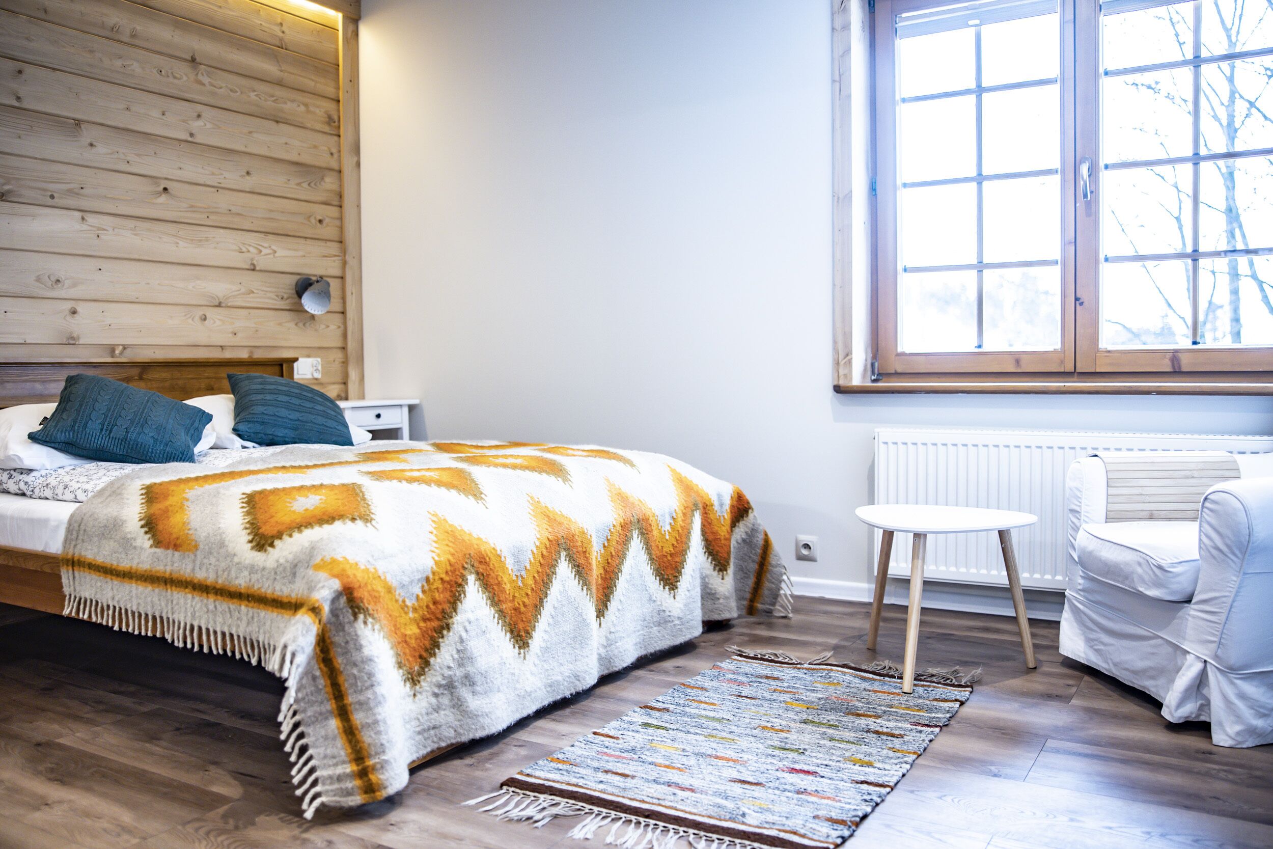 A bedroom with a wooden headboard, a bed with a patterned blanket, a small white table, and a white armchair.