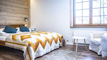 A bedroom with a wooden headboard, a bed with a patterned blanket, a small white table, and a white armchair.