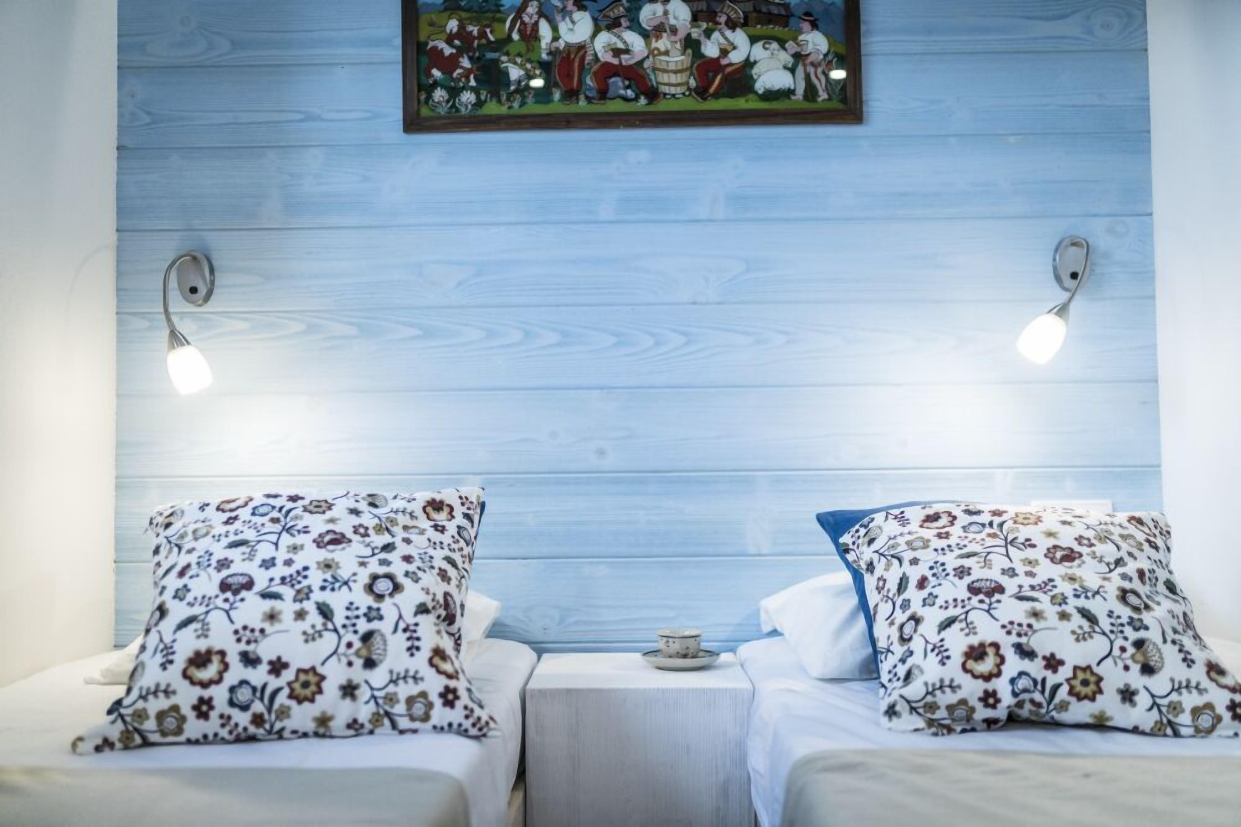Une chambre avec deux lits, chacun agrémenté d’oreillers à motifs floraux, une petite table avec une tasse, et un tableau accroché au mur au-dessus.