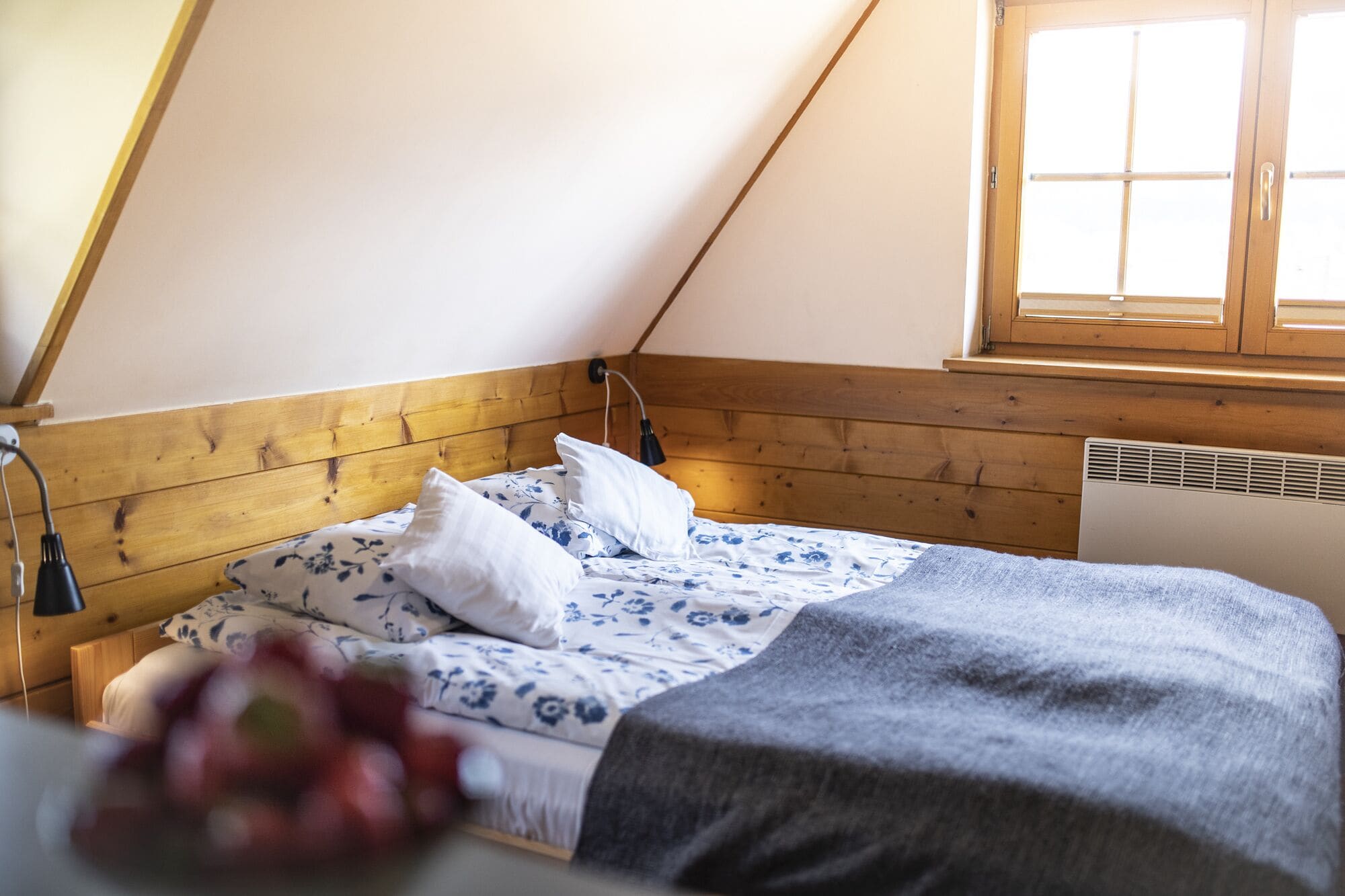 Quarto com teto inclinado, cabeceira de madeira, janela com persianas, cama com roupa de cama florida e luminária de cabeceira.