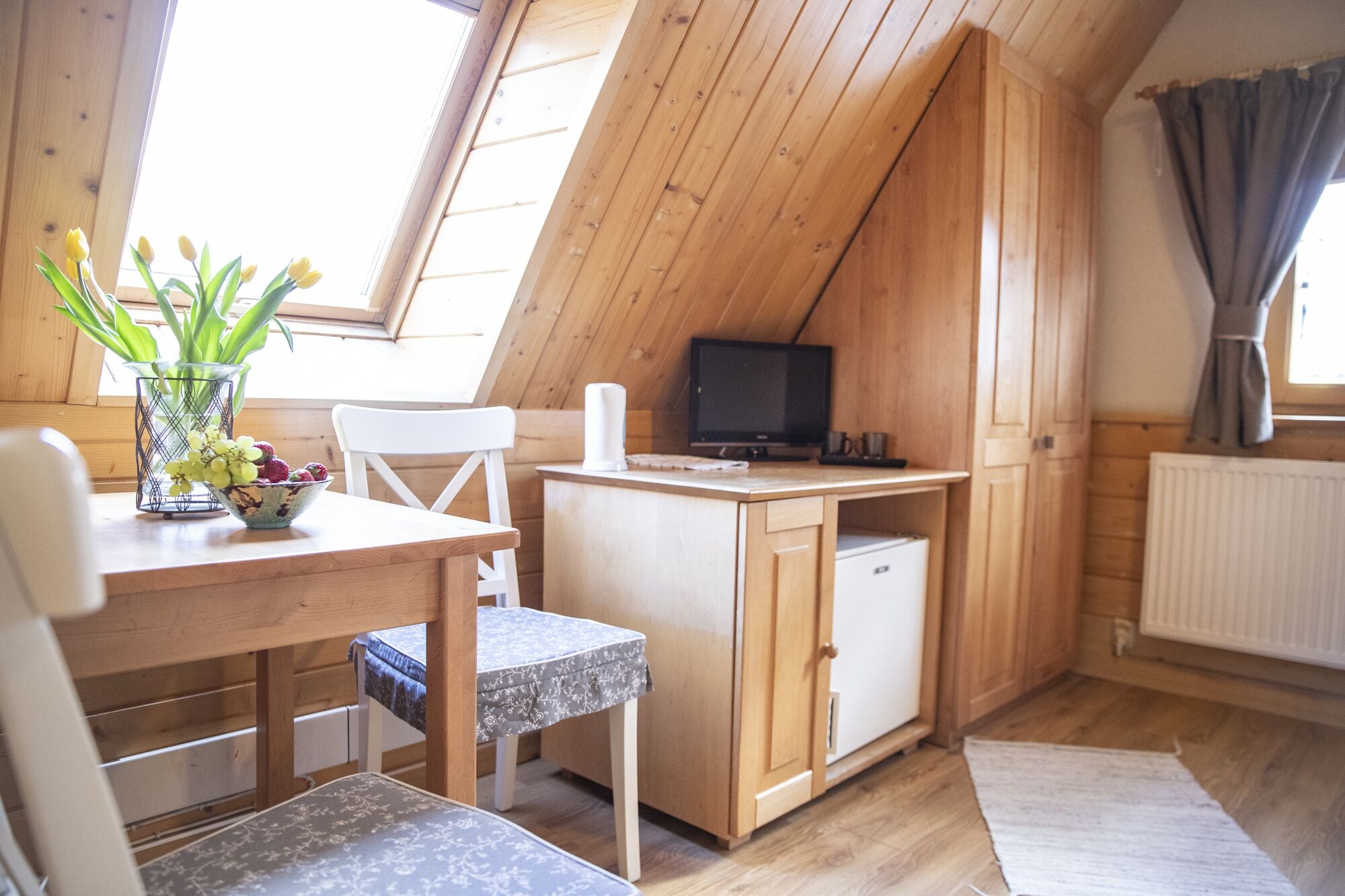 Een knus, houten kamer met een kleine keukenhoek, een eettafel met stoelen, een televisie en een raam met gordijnen.