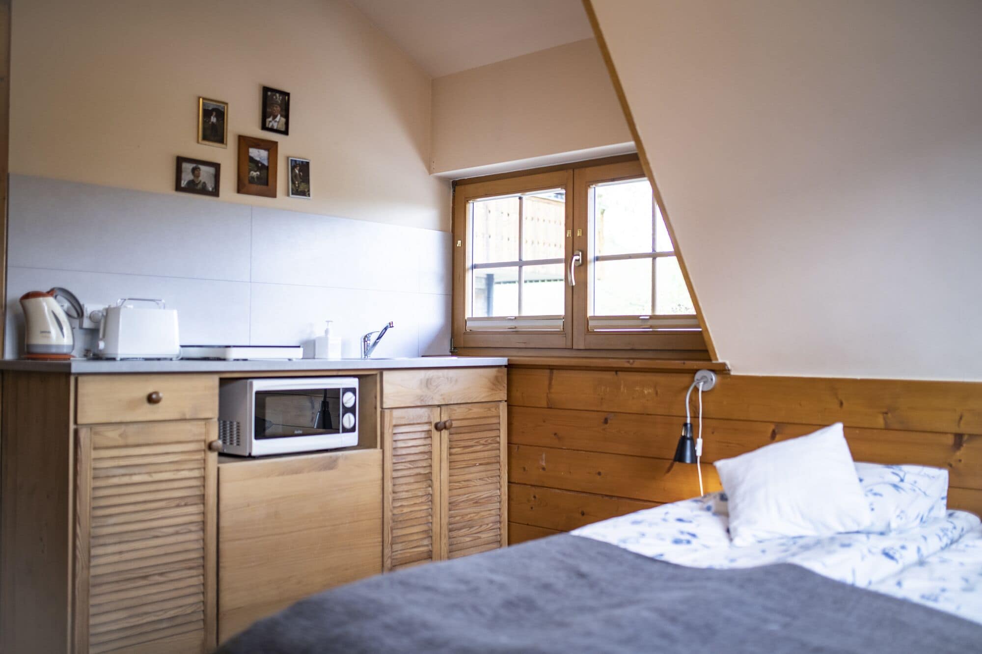A small, cozy room with a kitchenette, a bed with a floral duvet, a window with wooden shutters, and a sloped ceiling.