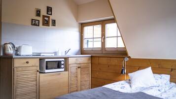 A small, cozy room with a kitchenette, a bed with a floral duvet, a window with wooden shutters, and a sloped ceiling.