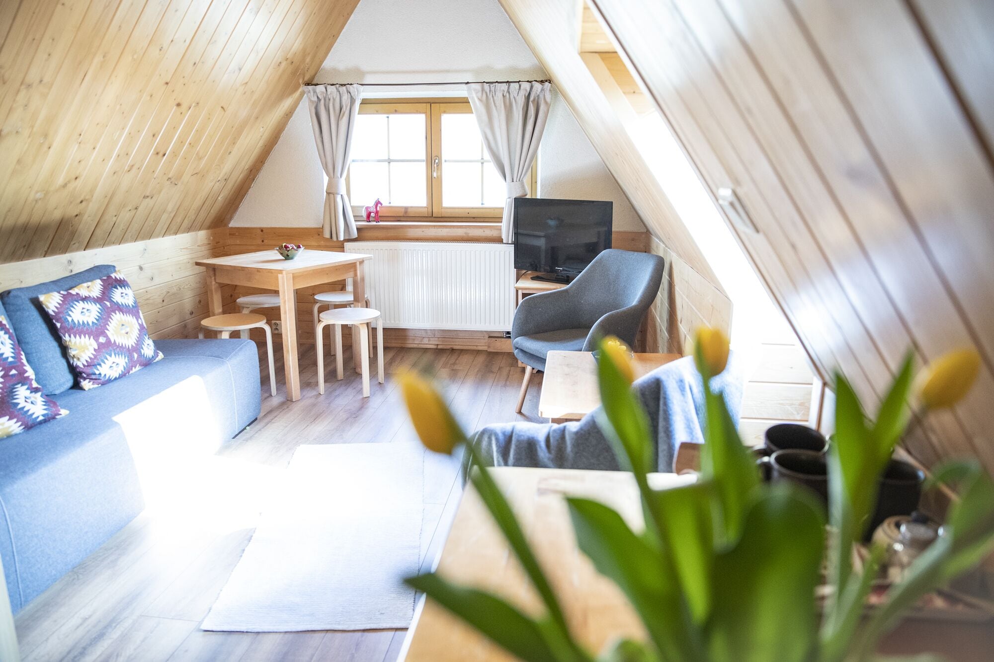 A cozy attic room with a wooden sloped ceiling, a sofa, a dining table with chairs, a TV, and a window with curtains.