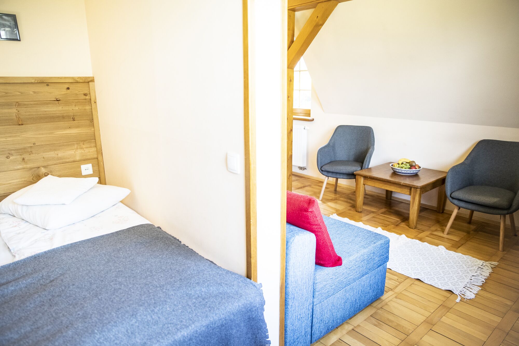 Una habitación con una cama, un sofá azul, dos sillas, una mesita con un frutero, y suelo de madera.