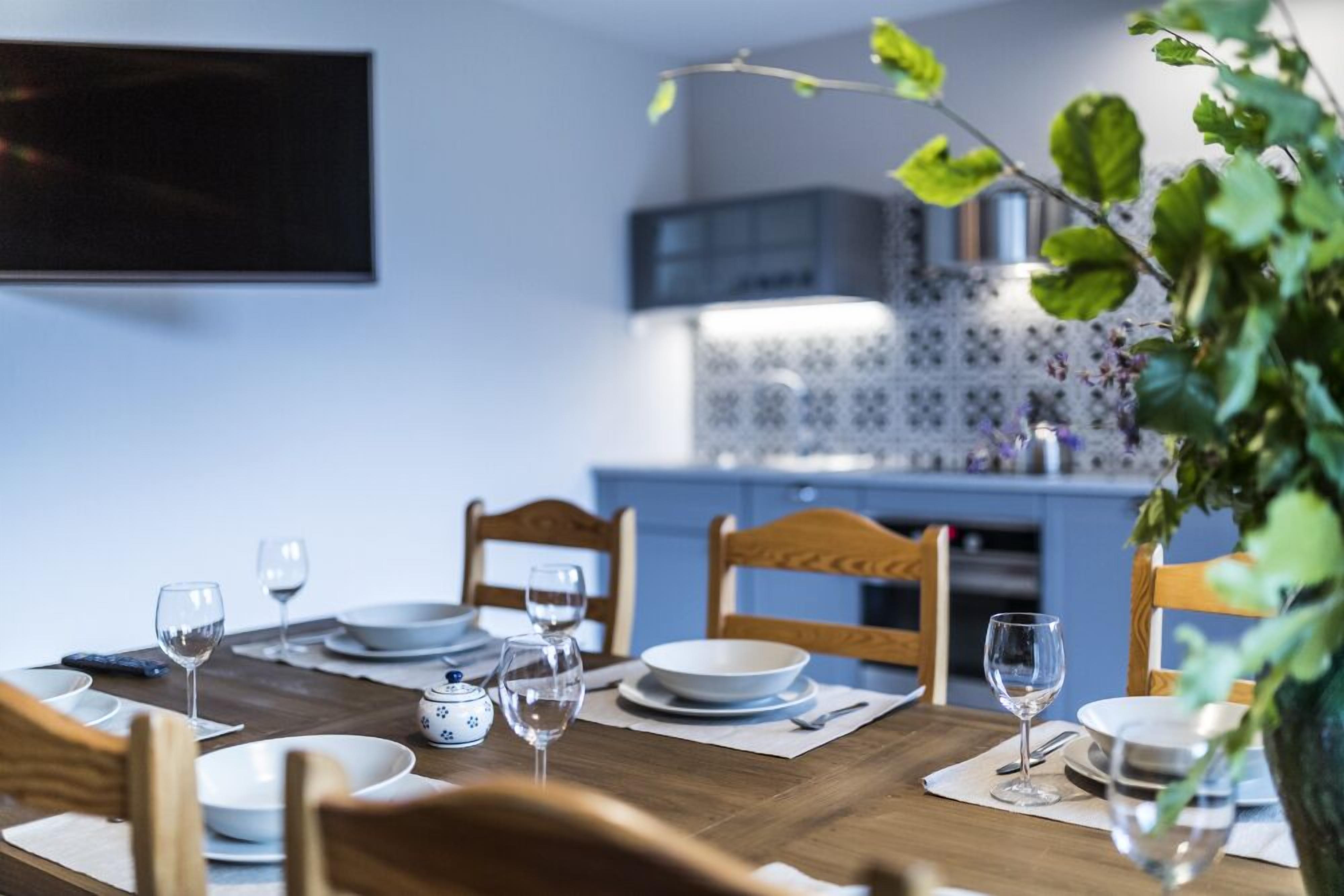 A dining table set with plates, glasses, and a teapot in a modern kitchen with blue cabinets and a tiled backsplash.