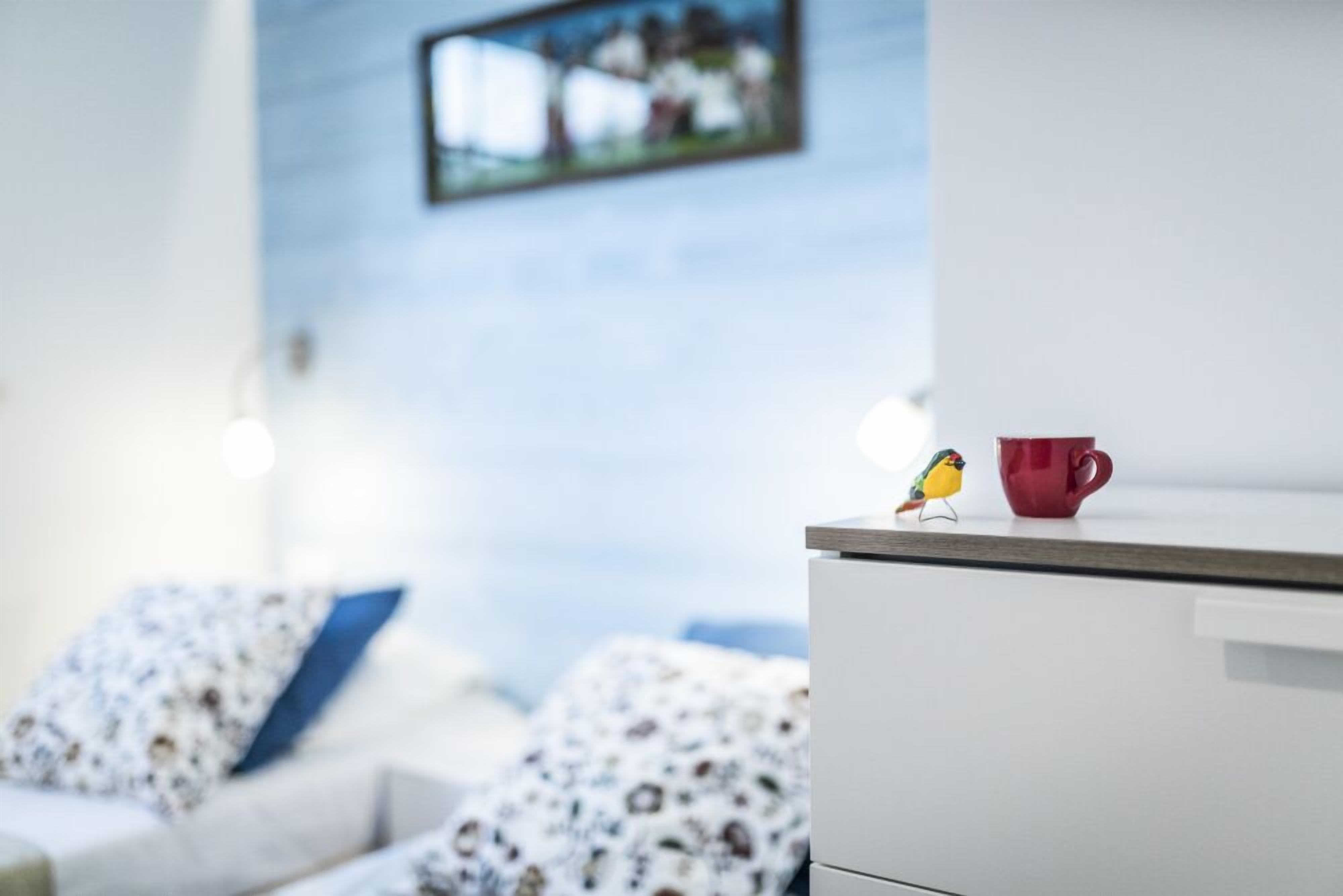 Ein modernes Zimmer mit einem weißen Schreibtisch, einer roten Tasse und einer kleinen Vogelstatue. An der Wand hängt ein gerahmtes Bild und auf dem Bett liegt ein Kissen mit Muster.