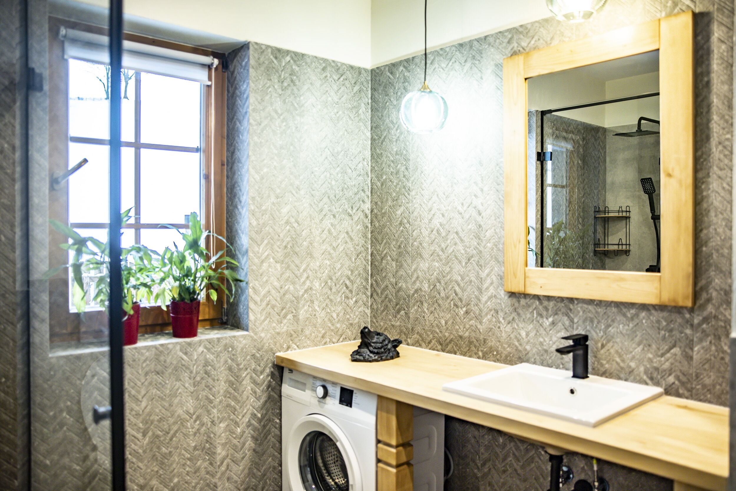 A compact bathroom with a washing machine, a sink, a mirror, and a window with potted plants.