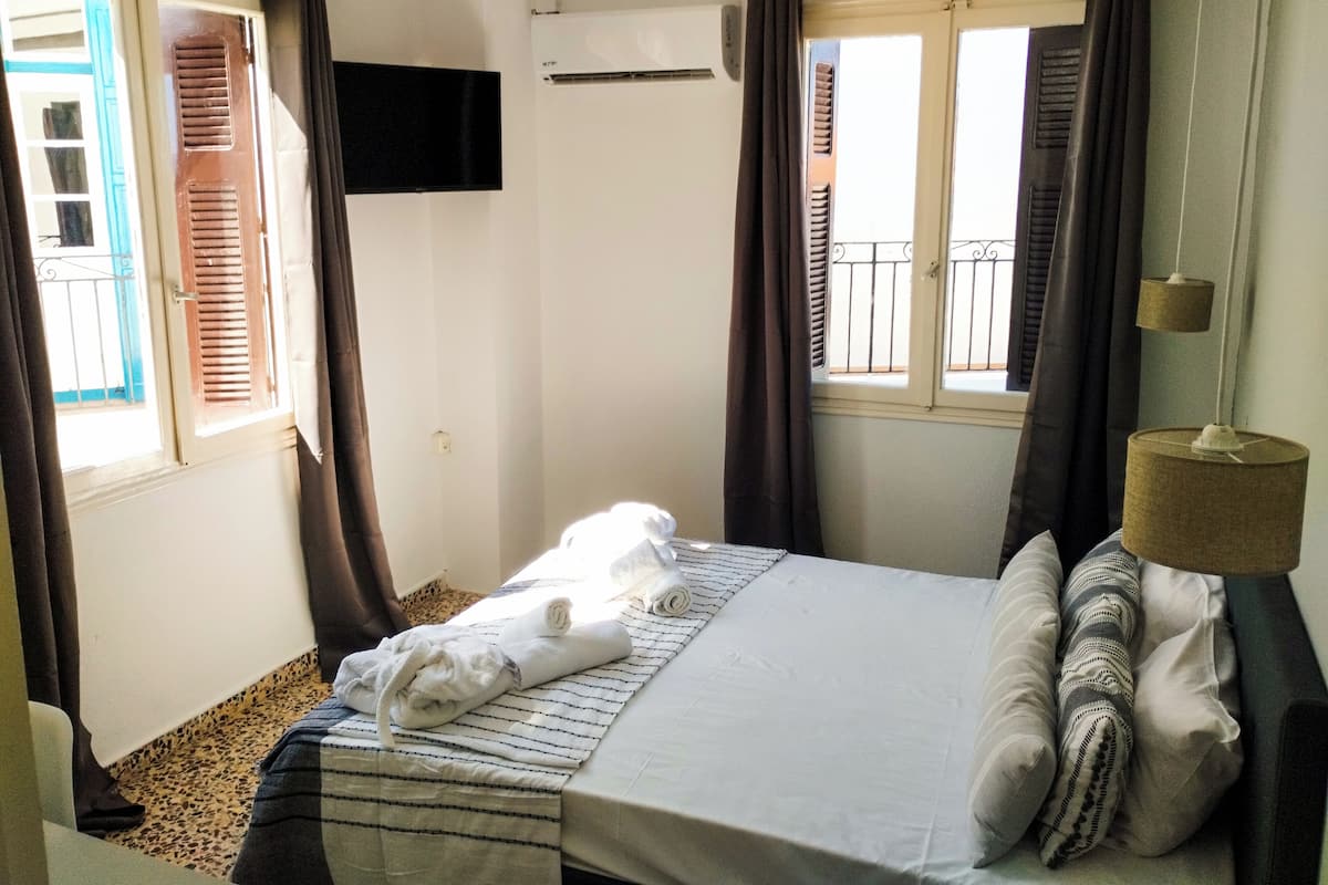 A bedroom with a bed, a window with curtains, a wall-mounted air conditioner, and a small table with a lamp.