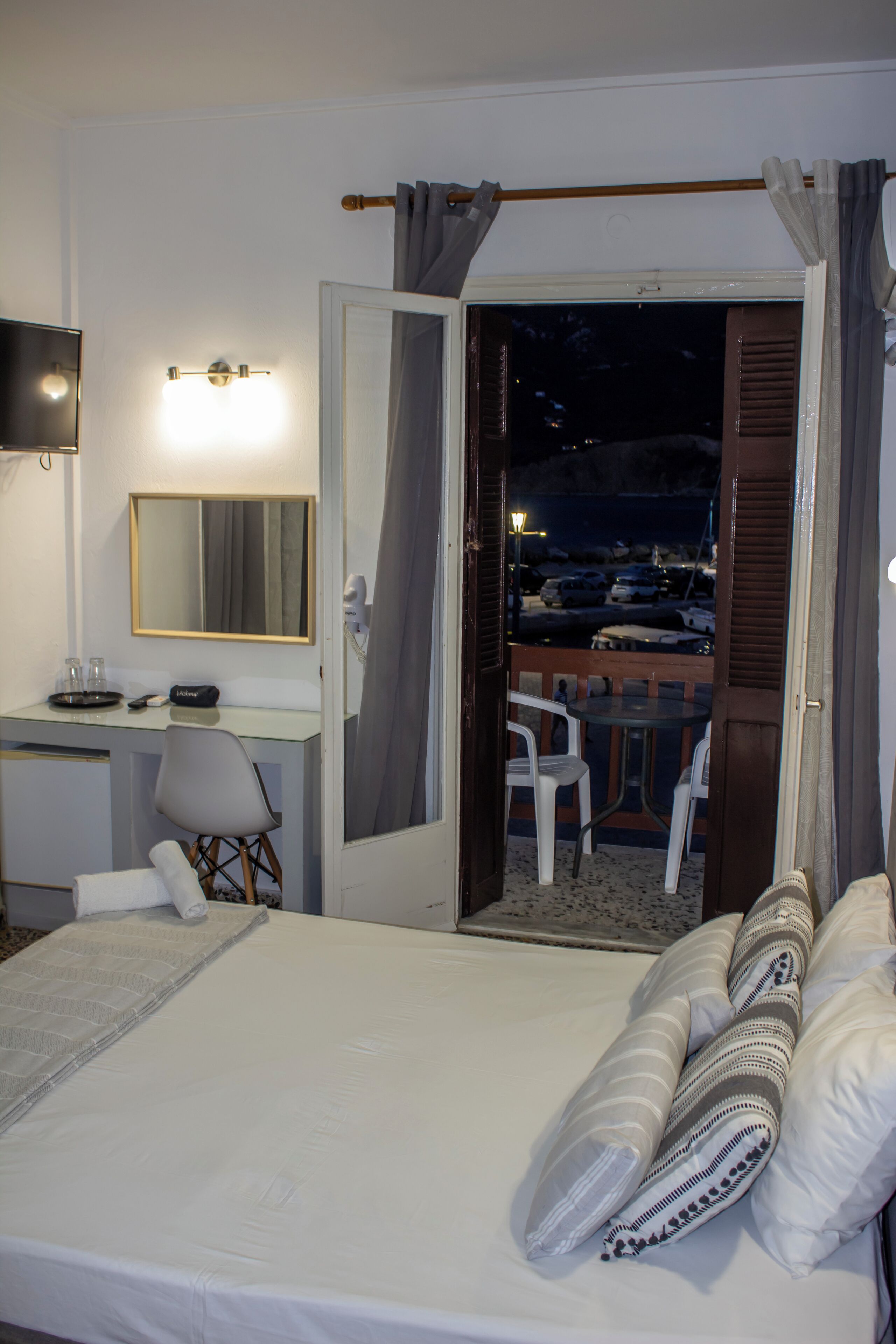 A hotel room with a bed, a desk, a chair, a TV, and a balcony with a view of the city.