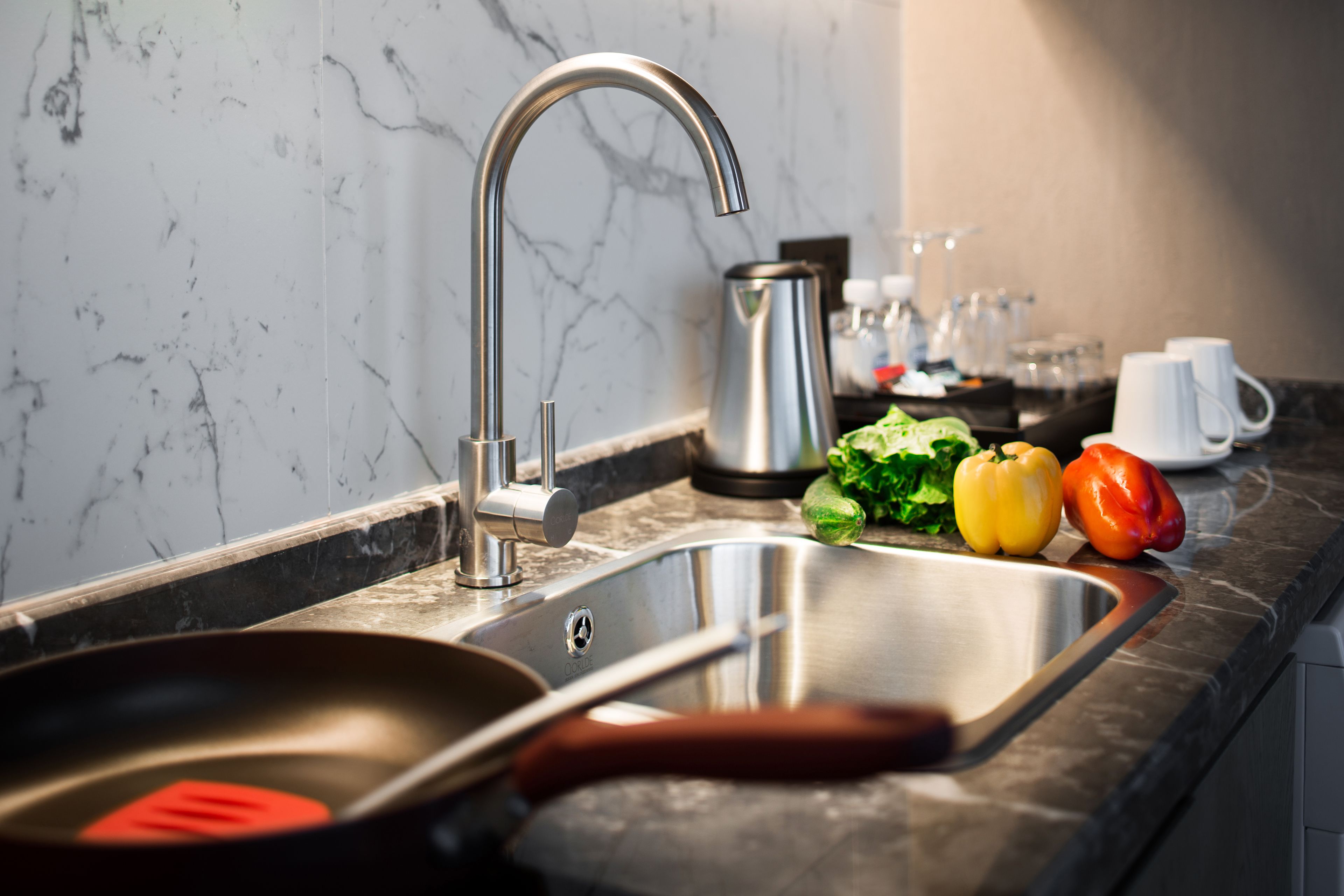A kitchen with a stainless steel sink, a kettle, and fresh vegetables on the counter.
