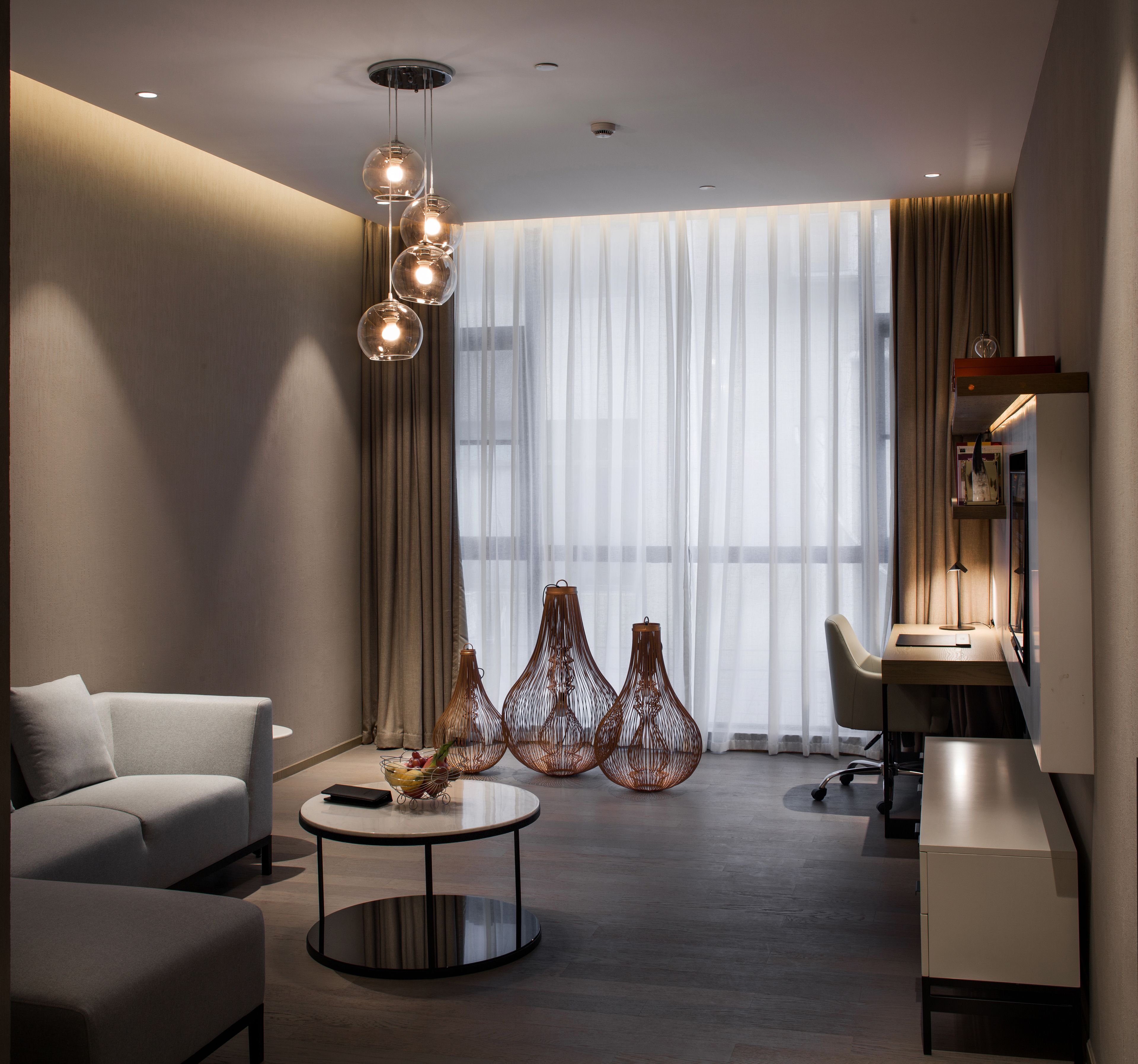 Una habitación de hotel moderna con sofá, una mesita redonda, escritorio y un ventanal con cortinas traslúcidas.