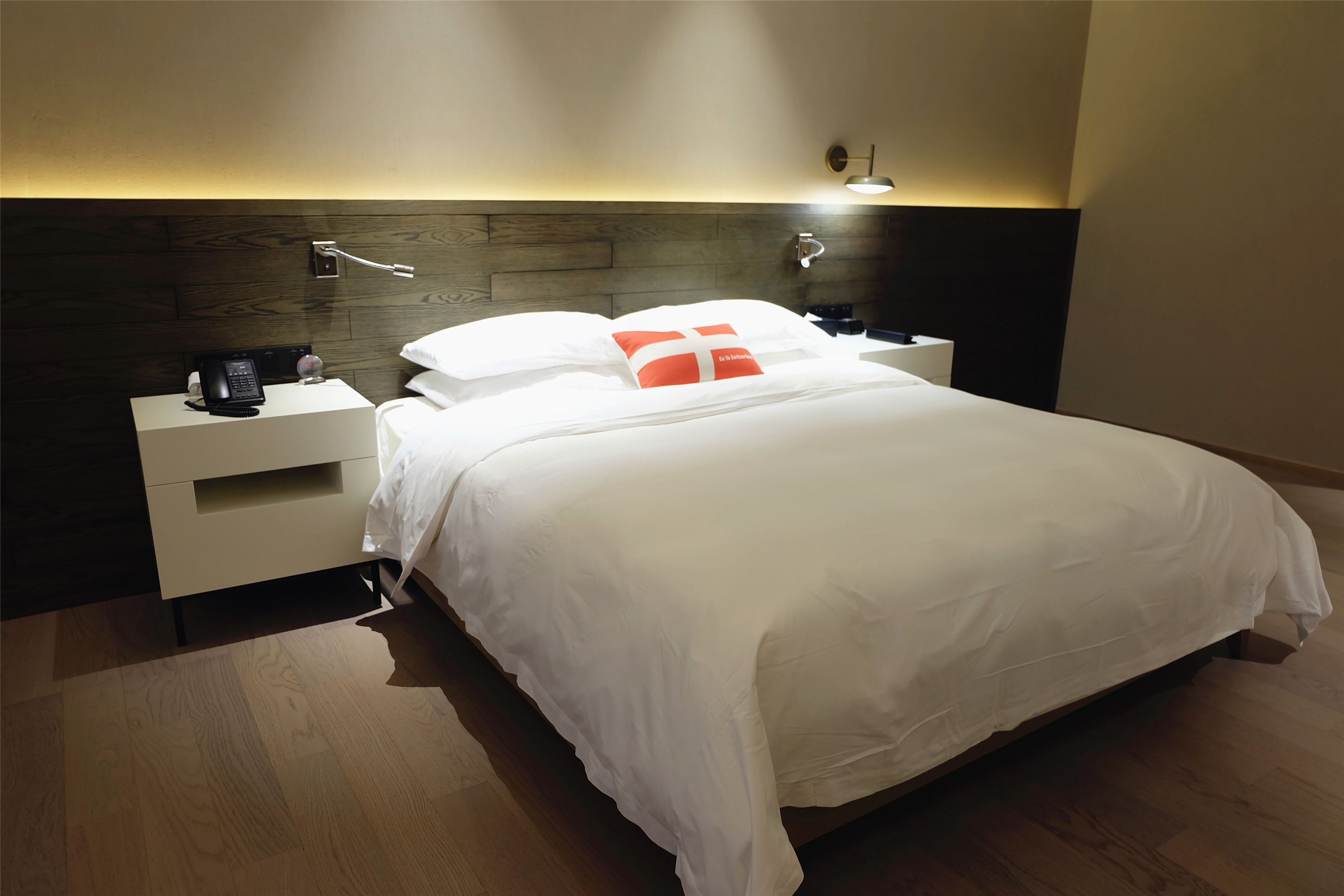Una cama bien hecha con ropa de cama blanca y un almohadón rojo, una mesita de noche con un teléfono y una lámpara, y un cabecero de madera.