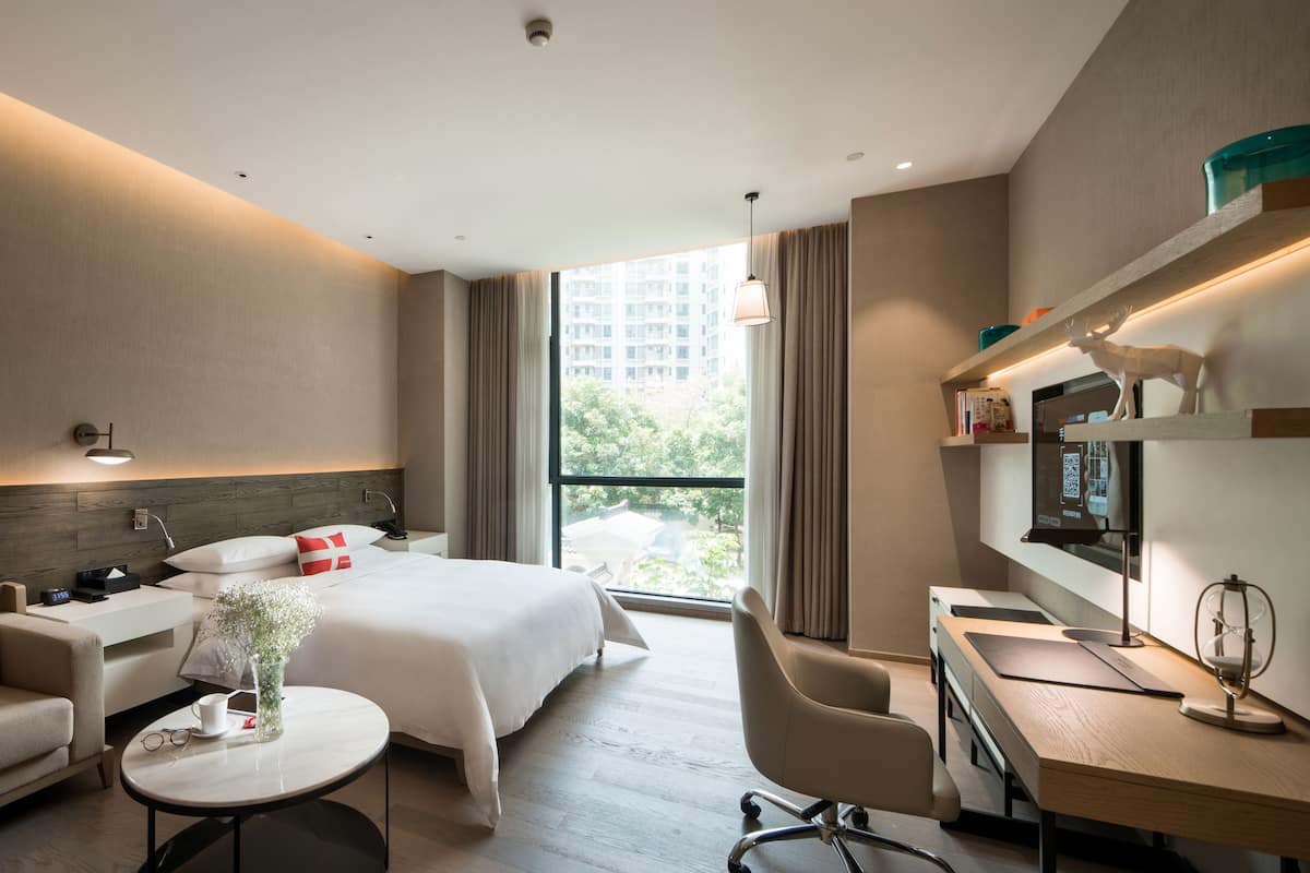 Ein modernes Hotelzimmer mit Bett, Sofa, Schreibtisch und Blick auf eine Stadtlandschaft.